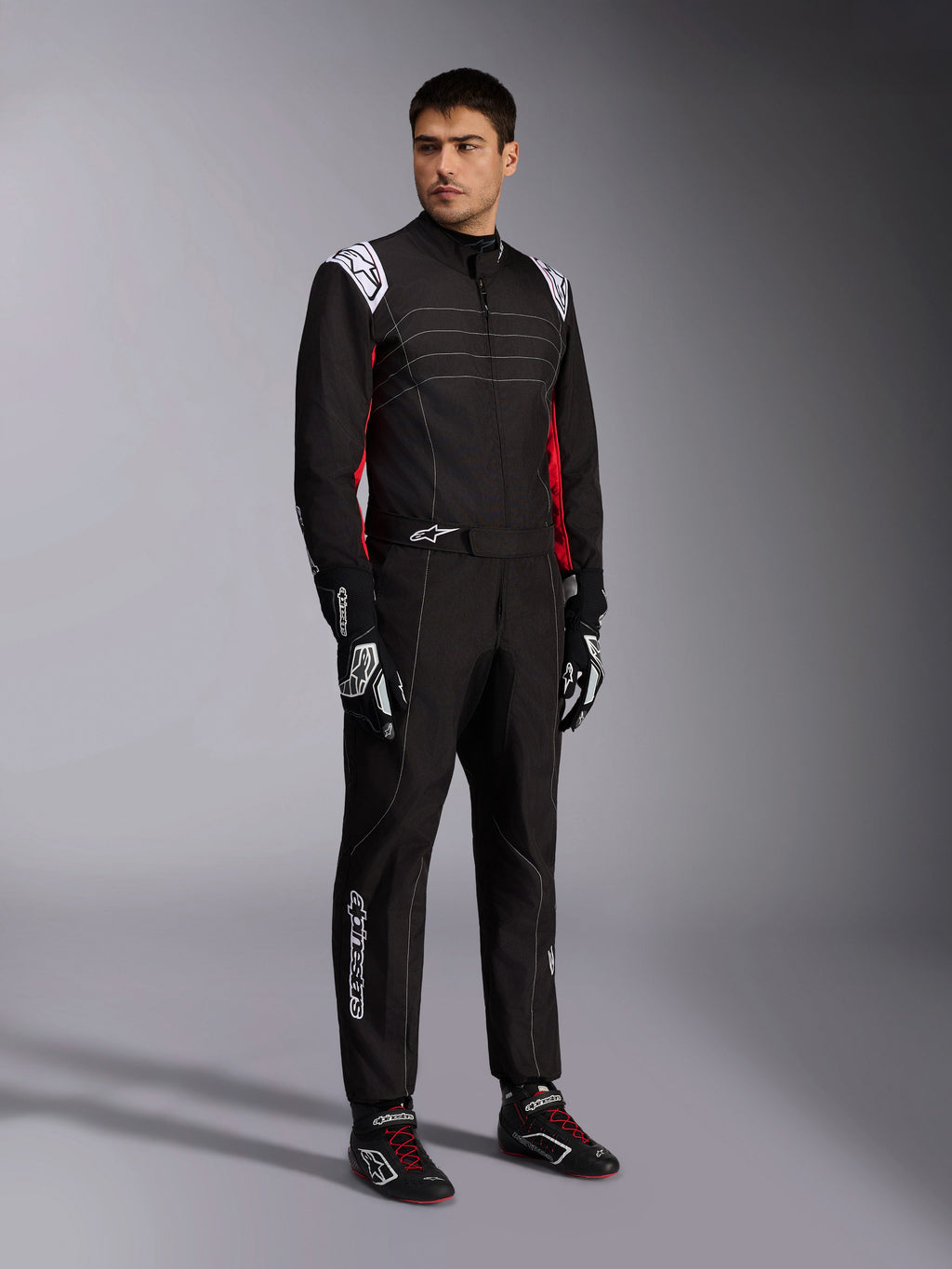Alpinestars KMX-9 V3 Anzug, Kart-Anzug in Schwarz, Rot und Weiß, getragen von einem Modell, mit weißen Epauletten und Logos, roten Farbakzenten an den Seiten, kombiniert mit passenden Handschuhen und Schuhen, professionelle Motorsportbekleidung.