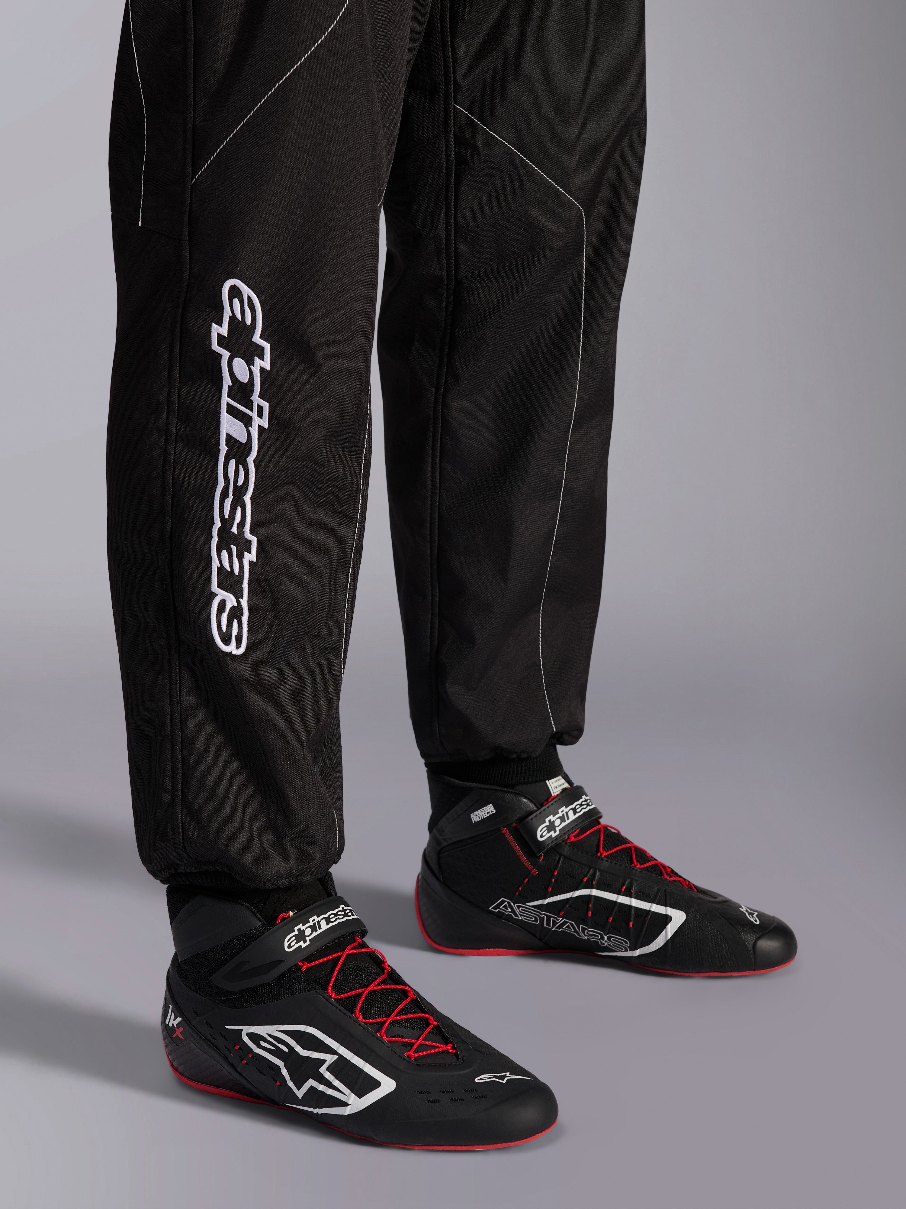 Alpinestars KMX-9 V3 Anzug, Kart-Anzug, Schwarz und Weiß, Detailansicht der Unterschenkel mit großem weißem Alpinestars-Branding, getragen mit schwarz-roten Rennstiefeln, professionelle Motorsport-Ausrüstung für den Kartsport