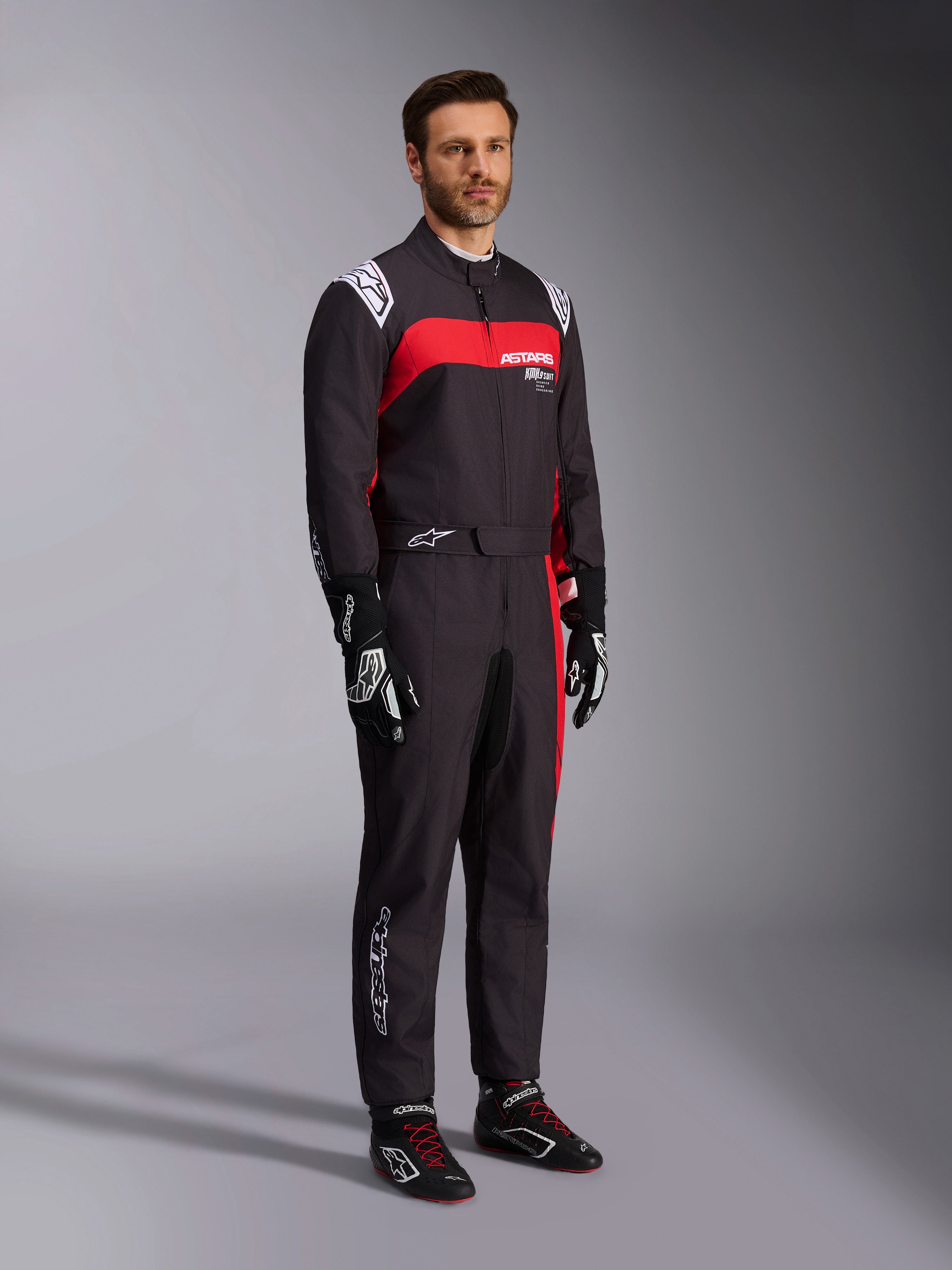 Male model wearing Alpinestars KMX-9 V3 Graphic 3 Suit, Karting Suit, Black and Red, featuring white shoulder epaulettes and red chest branding, paired with black racing gloves and shoes with red laces, professional motorsport apparel.