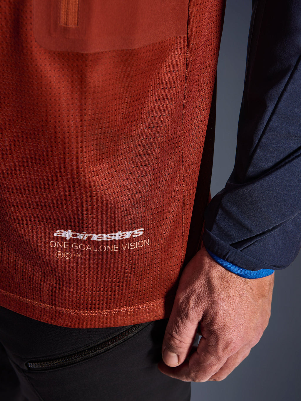 Alpinestars Maxdura Dual Trikot, Adventure-Motorradtrikot, Rostbraun und Mood Indigo, Nahaufnahme des Mesh-Torso-Gewebes und des navyfarbenen gewebten Ärmels, weißes Alpinestars „One Goal One Vision“-Branding am unteren Saum.