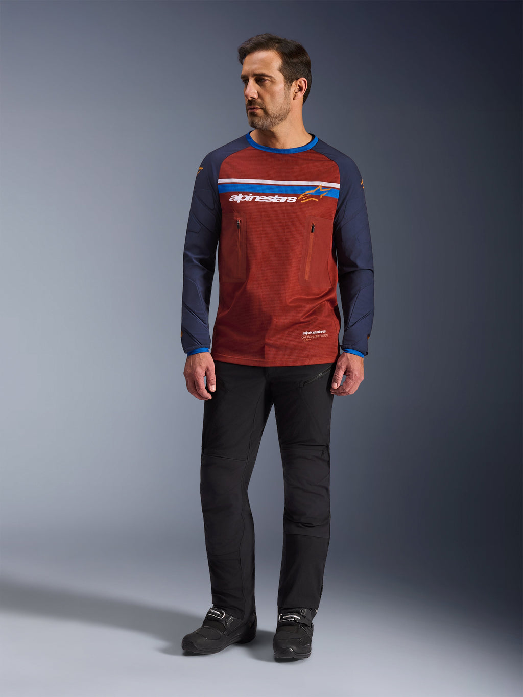 Alpinestars Maxdura Dual Jersey, Rostbraun und Mood Indigo, Adventure-Motorradtrikot mit Mesh-Torso und dunkelblauen Ärmeln, getragen von einem Model mit schwarzer Fahrhose und schwarzen Stiefeln, technische Dual-Sport-Bekleidung für Offroad-Fahrten