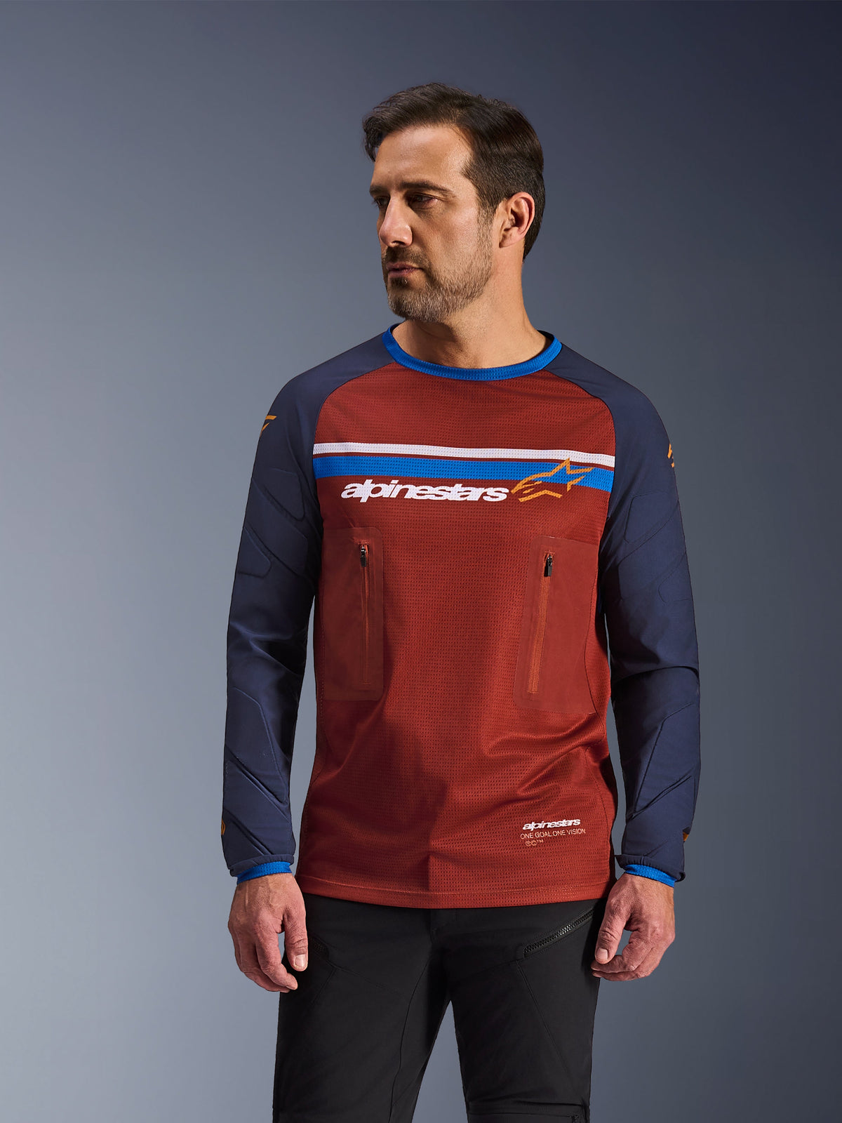 Männliches Model trägt Alpinestars Maxdura Dual Jersey, Adventure-Motorradtrikot in Rostbraun und Marineblau, mit zwei Brusttaschen mit Reißverschluss, horizontalem Bruststreifen mit Branding, technischem Mesh-Torso und strapazierfähigen gewebten Ärmeln.