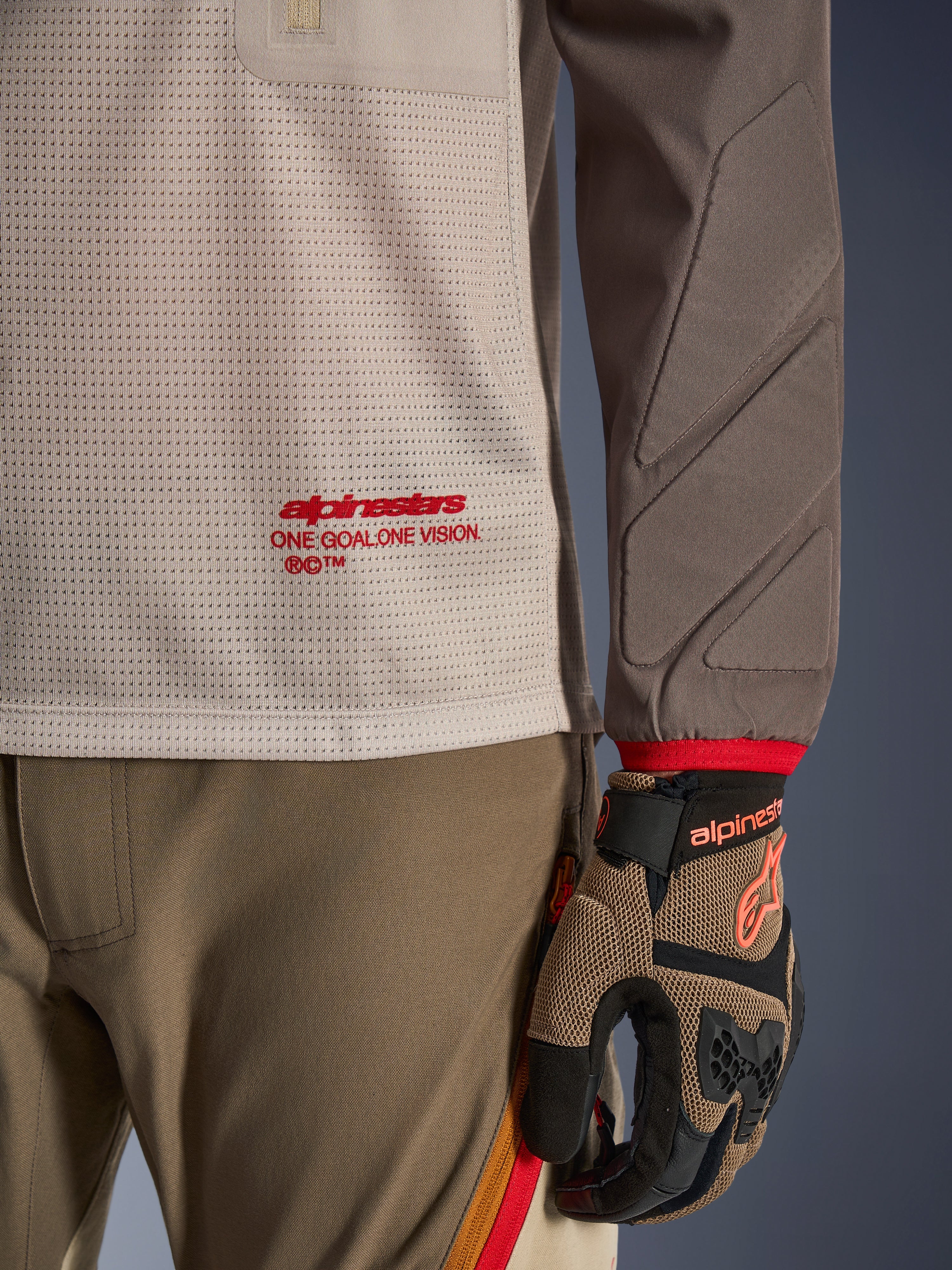Alpinestars Maxdura Dual Jersey in Aluminum Walnut, Detailansicht des belüfteten Mesh-Torsos mit rotem Branding, abriebfester Ärmel mit integrierter Polsterung, getragen mit passender Adventure-Fahrhose und Handschuhen