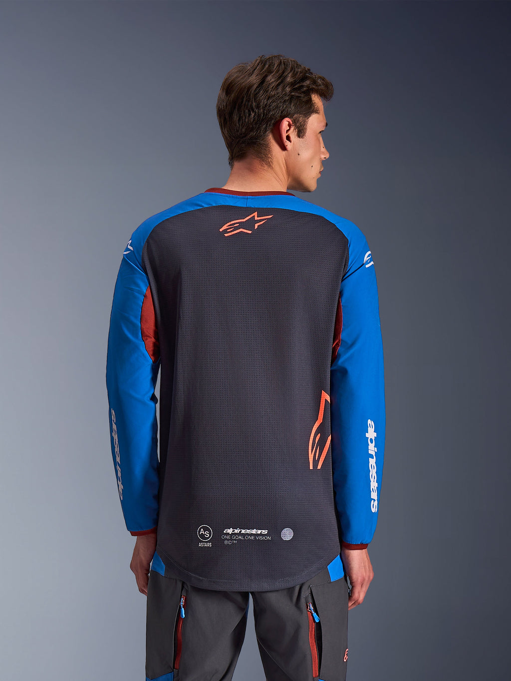 Alpinestars Maxdura Dual Jersey in Dunkelgrau und Royalblau, Rückansicht eines männlichen Modells, Mesh-Torso-Konstruktion mit orangefarbenem Astars-Logo, lange Ärmel mit weißem Branding, weinroter Kragen und Bündchen, getragen mit grauer technischer Hose, Dual-Sport- und Adventure-Motorradausrüstung