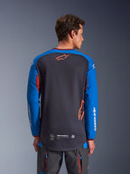 Alpinestars Maxdura Dual Jersey in Dunkelgrau und Royalblau, Rückansicht eines männlichen Modells, Mesh-Torso-Konstruktion mit orangefarbenem Astars-Logo, lange Ärmel mit weißem Branding, weinroter Kragen und Bündchen, getragen mit grauer technischer Hose, Dual-Sport- und Adventure-Motorradausrüstung