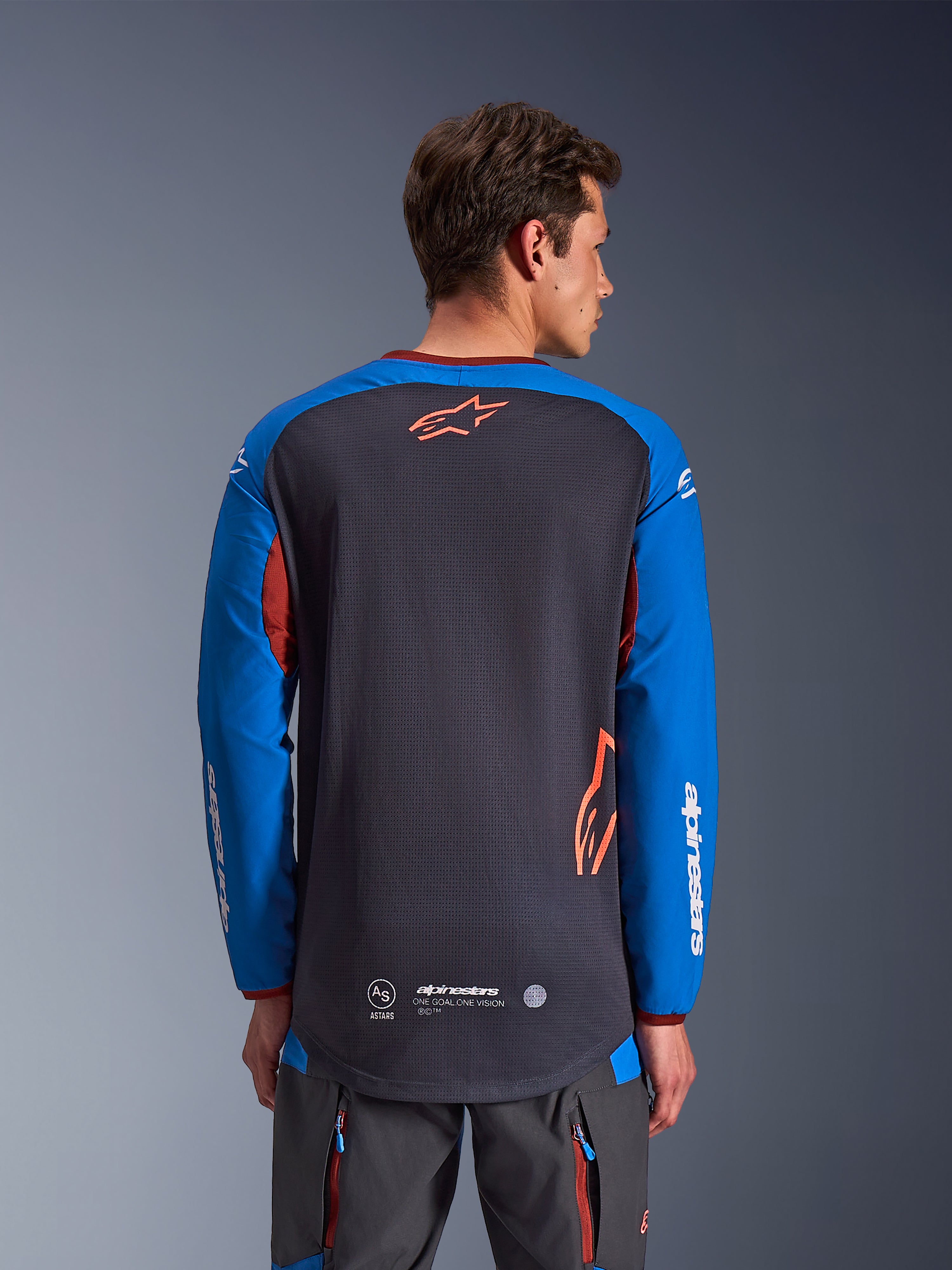 Alpinestars Maxdura Dual Jersey in Dunkelgrau und Royalblau, Rückansicht eines männlichen Modells, Mesh-Torso-Konstruktion mit orangefarbenem Astars-Logo, lange Ärmel mit weißem Branding, weinroter Kragen und Bündchen, getragen mit grauer technischer Hose, Dual-Sport- und Adventure-Motorradausrüstung