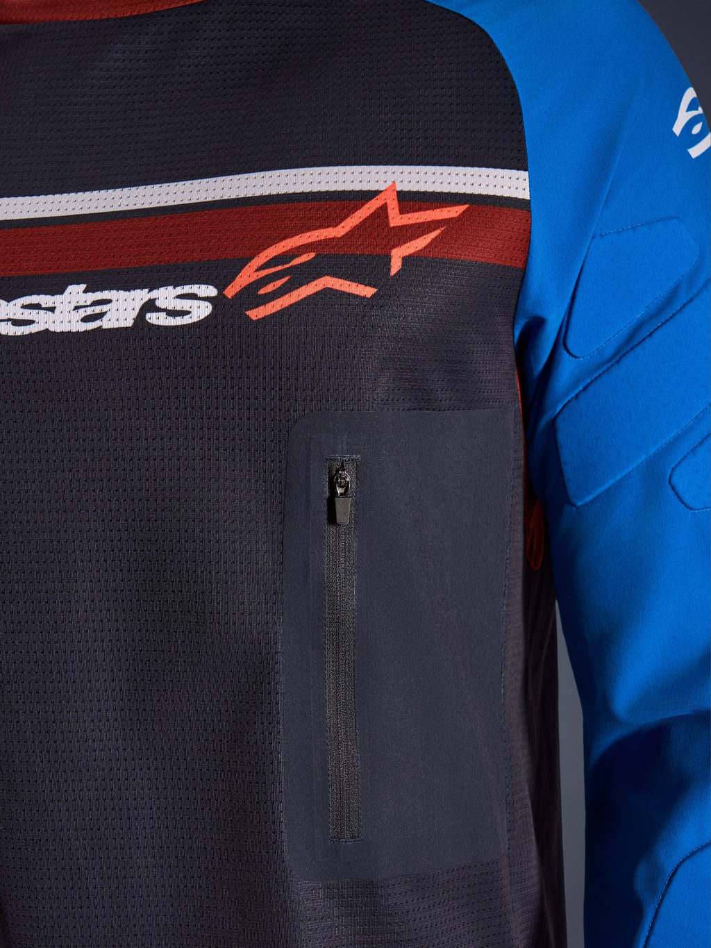 Alpinestars Maxdura Dual Jersey, Nahaufnahme von Brust und Ärmel, Dunkelgrau und Königsblau, mit Reißverschluss-Brusttasche, atmungsaktivem Mesh-Torso und integrierter Polsterung an den Ärmeln, Alpinestars-Logo auf roten und weißen Streifen.