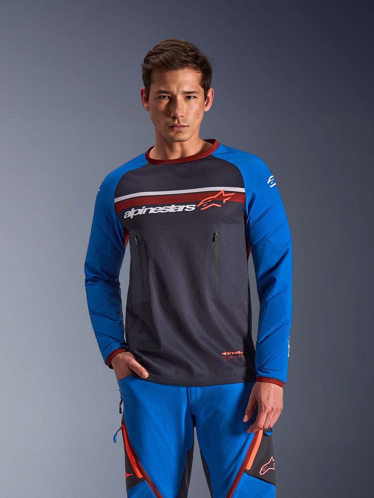 Mann im Alpinestars Maxdura Dual Jersey, Deep Gray und Royal Blue mit orangefarbenen Akzenten, Mesh-Torso und Brusttaschen mit Reißverschluss, kombiniert mit passender blauer Adventure-Fahrhose, technische Dual-Sport- und Offroad-Bekleidung