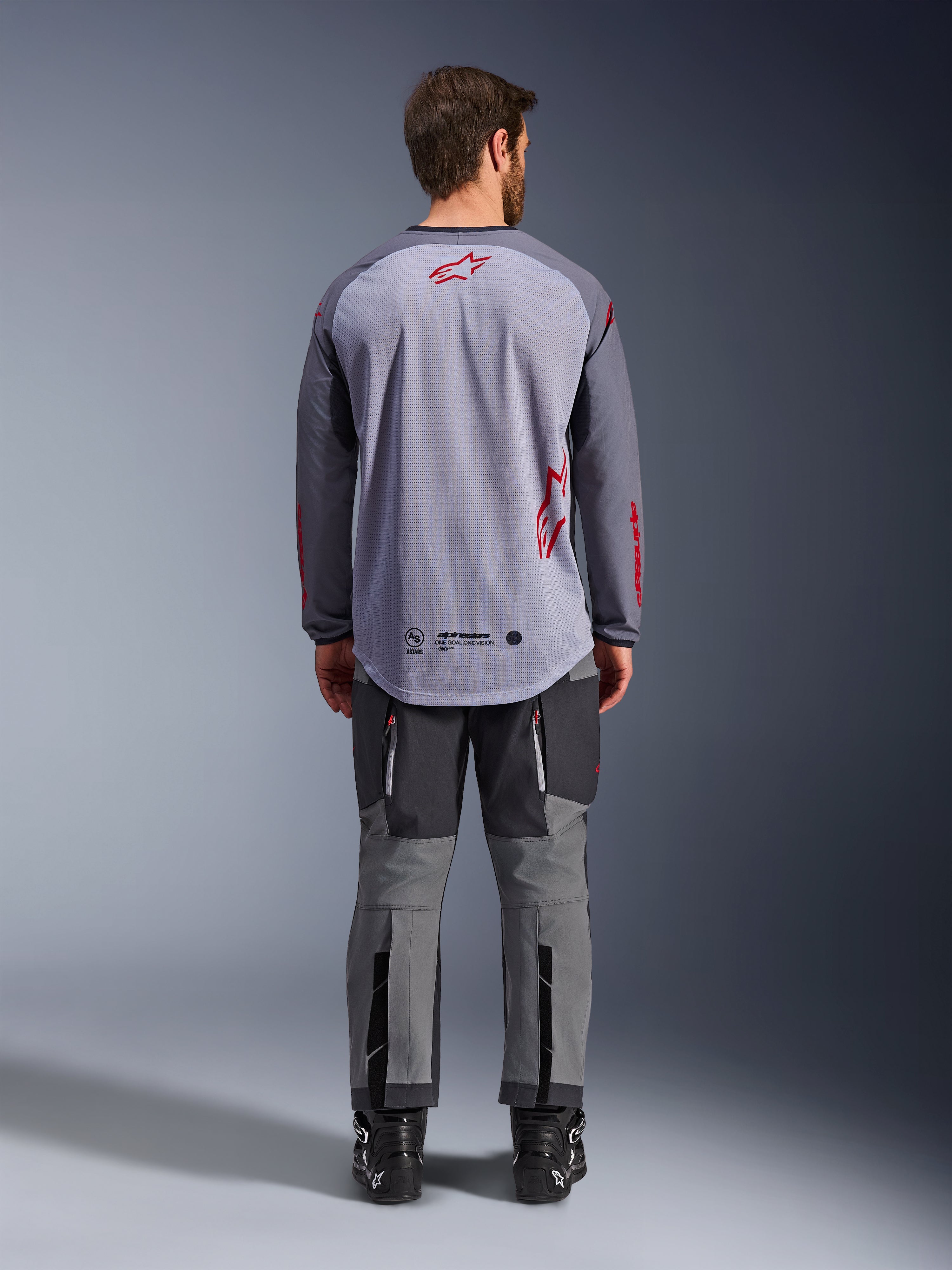 Männliches Model in der Rückansicht trägt das Alpinestars Maxdura Dual Jersey, Adventure-Trikot in Mittelgrau und Dunkelgrau mit roten Logo-Akzenten, Mesh-Rückenkonstruktion, kombiniert mit passender grauer Offroad-Hose und schwarzen Stiefeln