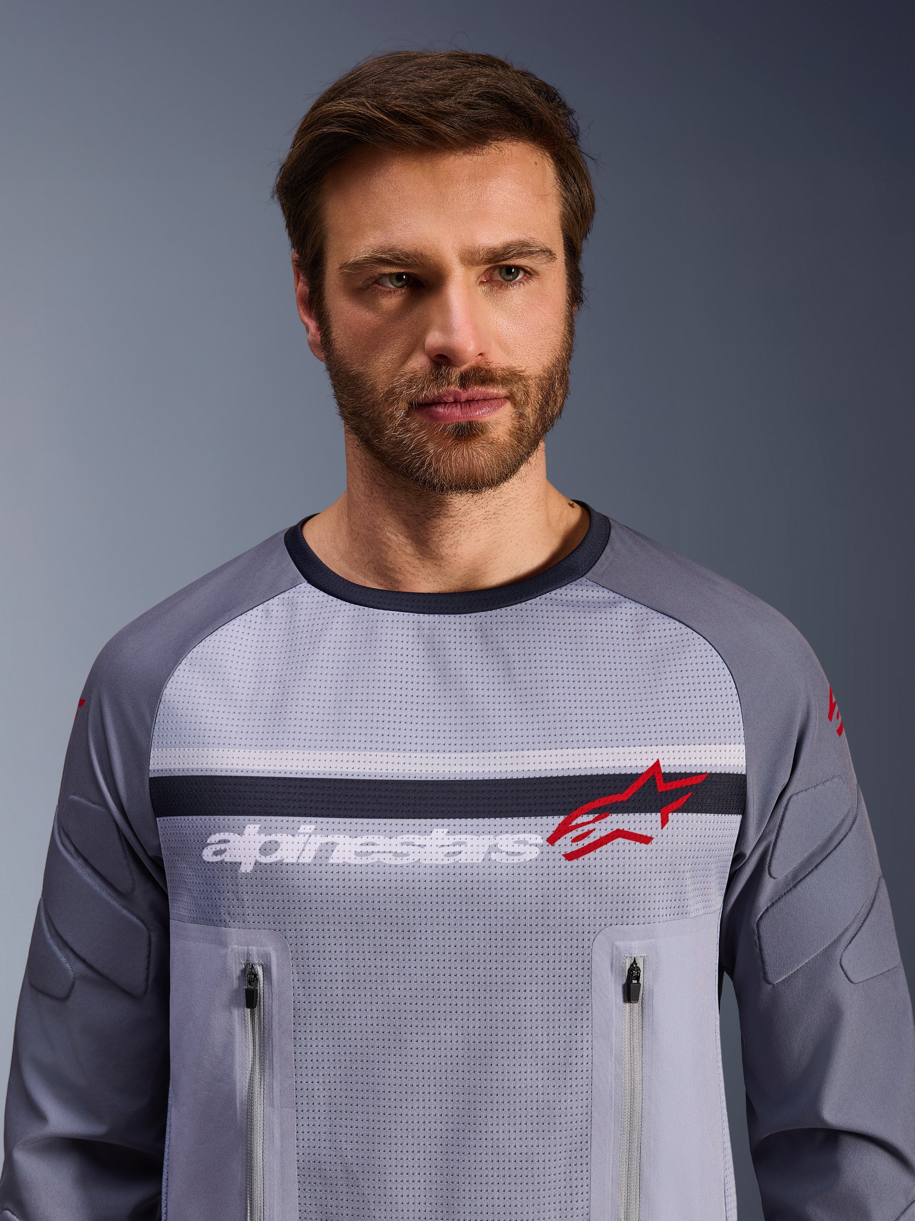 Alpinestars Maxdura Dual Jersey, Mittelgrau und Dunkelgrau, getragen von einem männlichen Model, Mesh-Torso für Belüftung, zwei Brusttaschen mit Reißverschluss, weißes „alpinestars“-Branding und rotes Stern-Logo, Performance-Offroad-Adventure-Ausrüstung