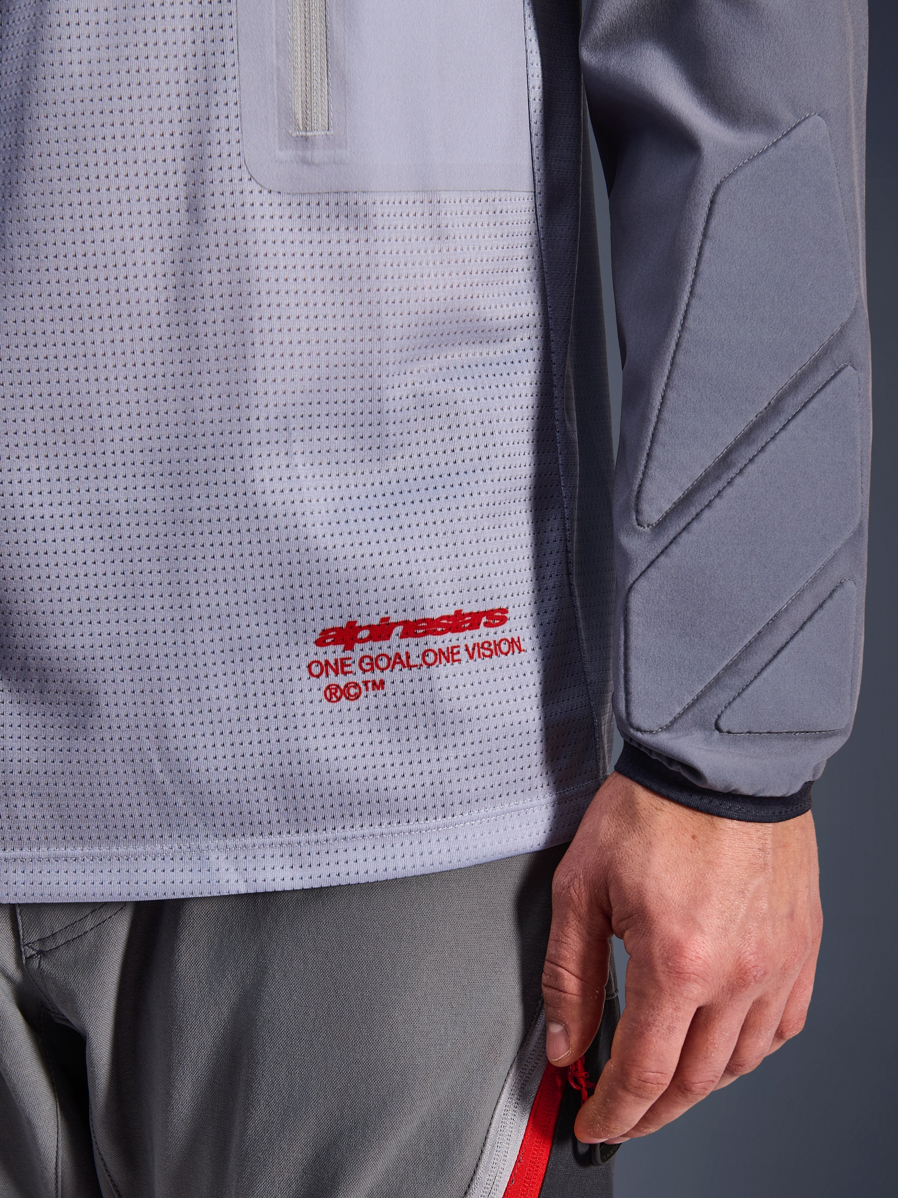 Alpinestars Maxdura Dual Jersey, Mid Gray und Dark Gray, Nahaufnahme des atmungsaktiven Mesh-Torsos mit rotem Branding, Reißverschlusstaschen-Detail und integrierter Polsterung am Ärmel, technische Adventure-Motorradausrüstung