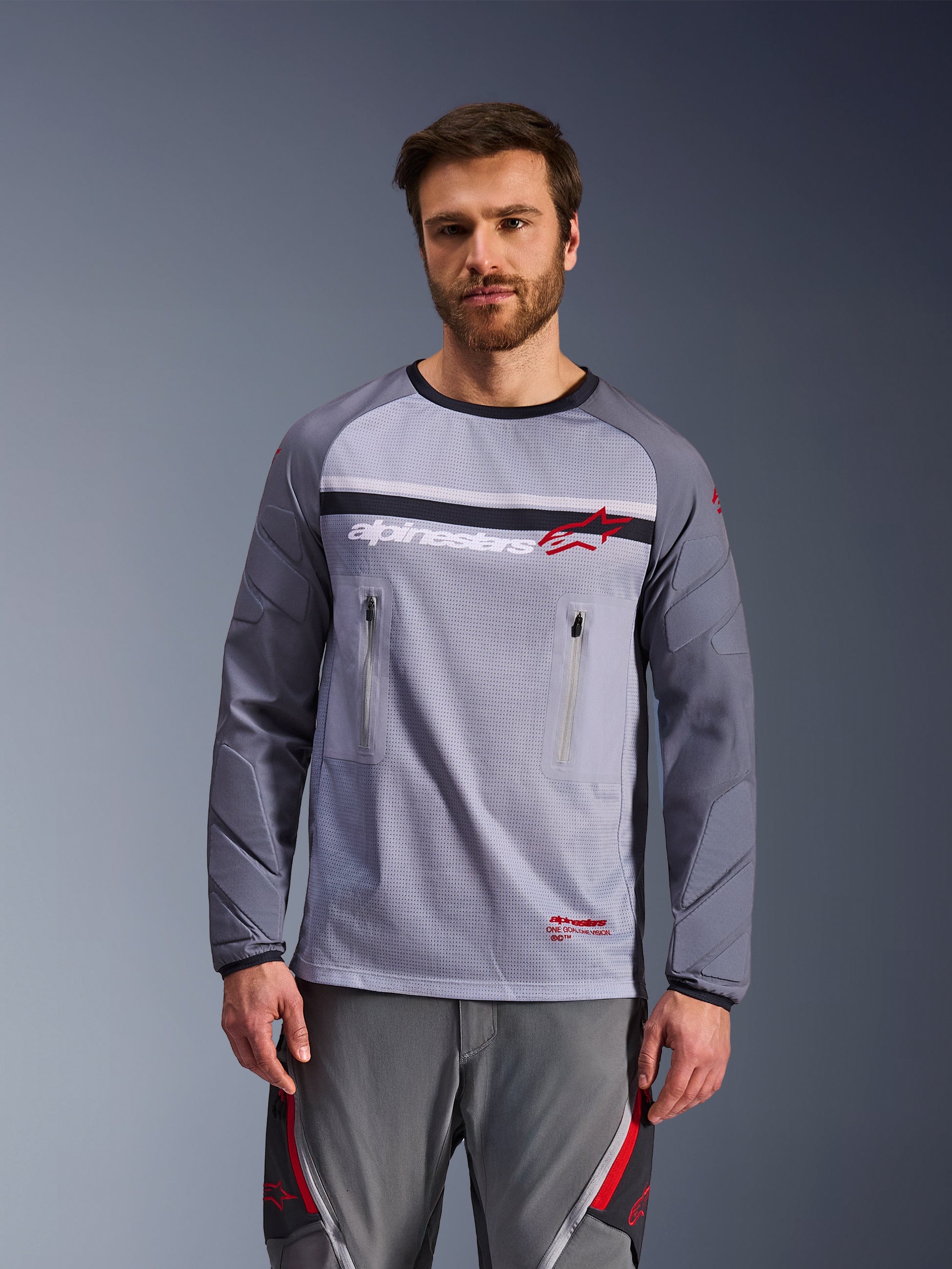 Alpinestars Maxdura Dual Jersey, Adventure-Motorrad-Trikot, Mittelgrau und Dunkelgrau, getragen von einem Model mit passender grauer Hose, mit Brusttaschen mit Reißverschluss, Mesh-Konstruktion am Torso und gepolsterten abriebfesten Ärmeln, Offroad-Ausrüstung