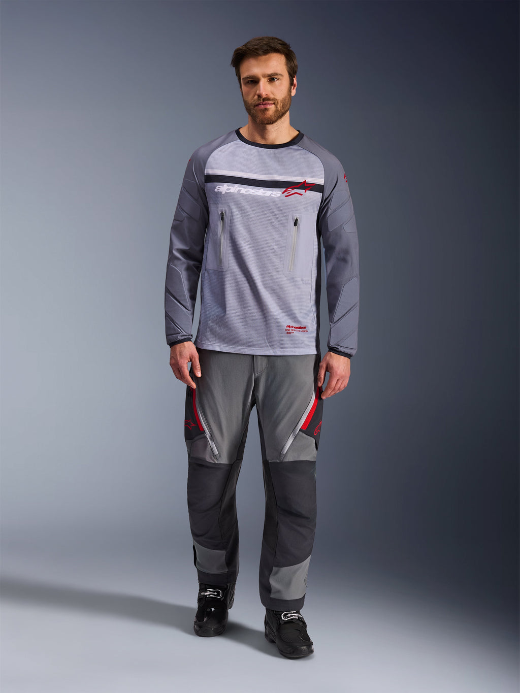 Alpinestars Maxdura Dual Jersey, Mittelgrau und Dunkelgrau, Adventure-Motorradtrikot mit Mesh-Torso und Brusttaschen mit Reißverschluss, abriebfeste Ärmel mit integrierter Polsterung, getragen mit passender grauer Performance-Hose mit roten Akzenten, technische Offroad-Fahrausrüstung