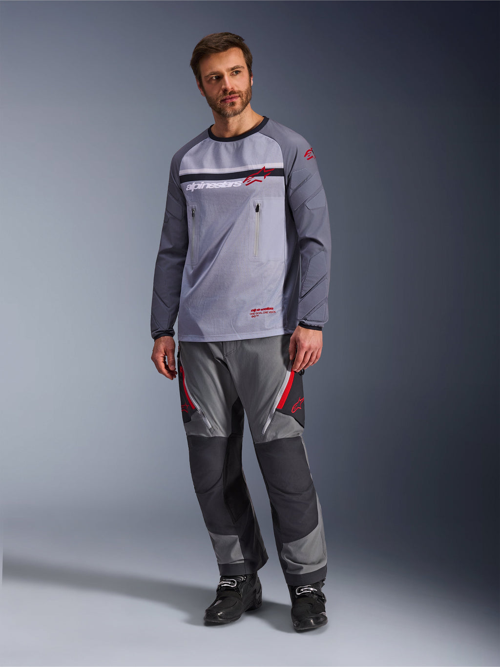 Ein Mann trägt das Alpinestars Maxdura Dual Jersey in Mittelgrau und Dunkelgrau, kombiniert mit einer farblich abgestimmten grau-schwarzen Adventure-Hose und schwarzen Stiefeln, mit atmungsaktivem Mesh-Torso, Brusttaschen mit Reißverschluss und roten Logo-Akzenten.