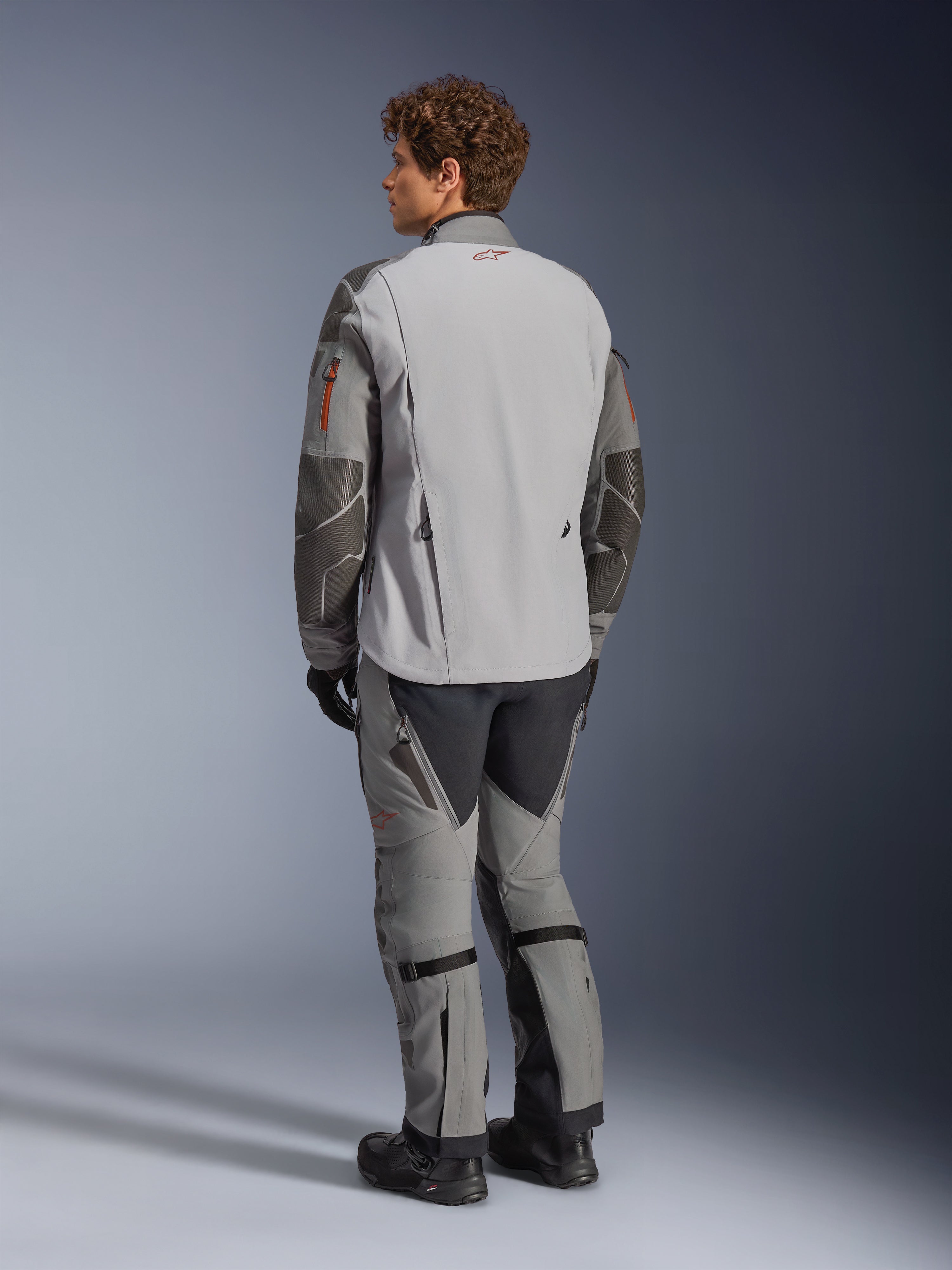 Alpinestars Nazca 3L GORE-TEX PRO Jacke, Adventure-Motorradjacke, Dunkelgrau und Mittelgrau, Rückansicht getragen mit passender Adventure-Hose und schwarzen Stiefeln, mit dunkelgrauen Superfabric-Verstärkungen an den Ärmeln, orangen Reißverschluss-Akzenten und einem kleinen roten Logo am rückseitigen Kragen.