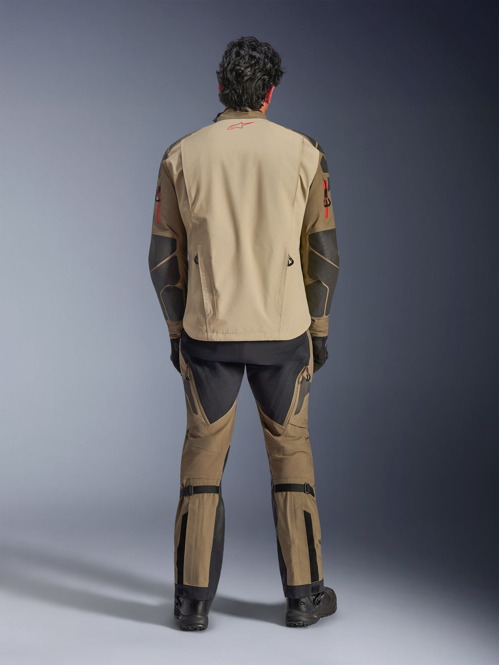 Alpinestars Nazca 3L GORE-TEX PRO Jacke in Walnut Rock Brown, Rückansicht des Fahrers, zeigt Superfabric-Verstärkungen an den Ellbogen und hintere Belüftungsreißverschlüsse, getragen mit passender Adventure-Hose, schwarzen Stiefeln und Handschuhen, robuste technische Fahrbekleidung für extreme Abenteuer.