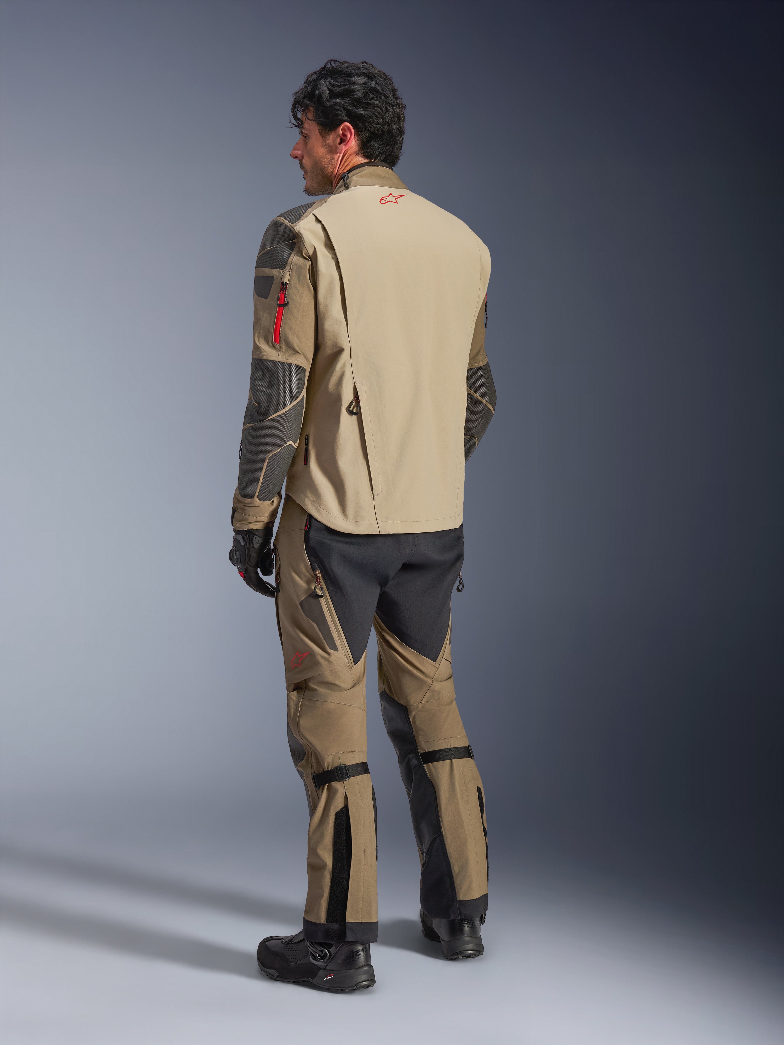 Alpinestars Nazca 3L GORE-TEX PRO Jacke, Abenteuer-Motorradjacke, Walnut Rock Brown, Rückansicht eines Fahrers in kompletter Abenteuer-Ausrüstung mit passender Hose und Stiefeln, beige und schwarze Paneele, rotes Logo am oberen Rücken, strapazierfähige Textilkonstruktion.