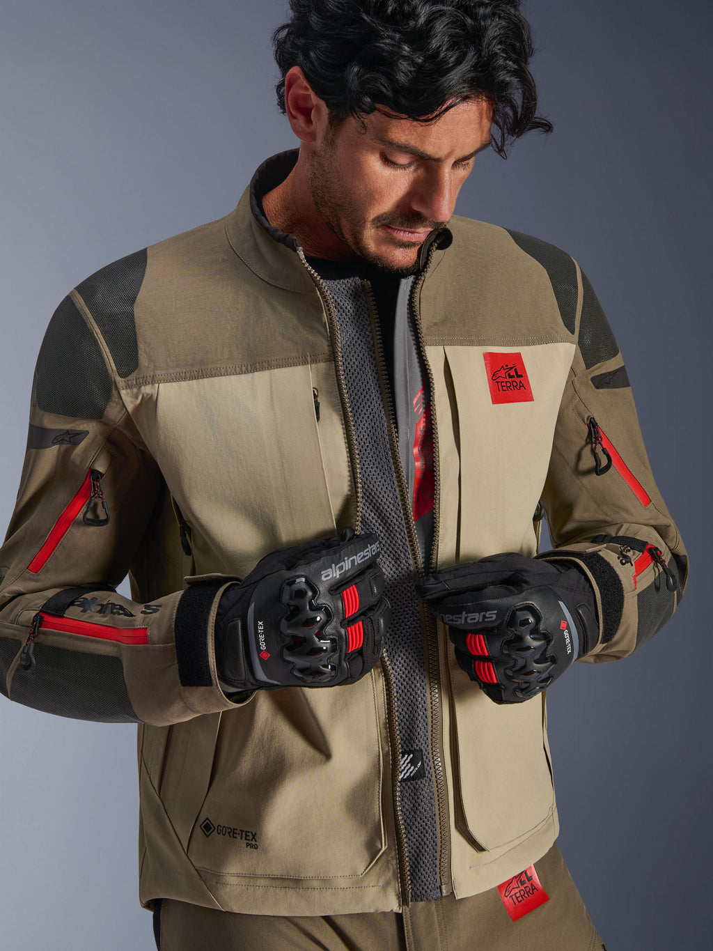 Alpinestars Nazca 3L GORE-TEX PRO Jacke, Walnut Rock Brown, Adventure-Motorradjacke mit offenem Frontreißverschluss und zentralem Mesh-Belüftungspanel, rotes All Terra-Branding, getragen mit schwarzen GORE-TEX-Motorradhandschuhen, technische Ausrüstung für Offroad-Touren