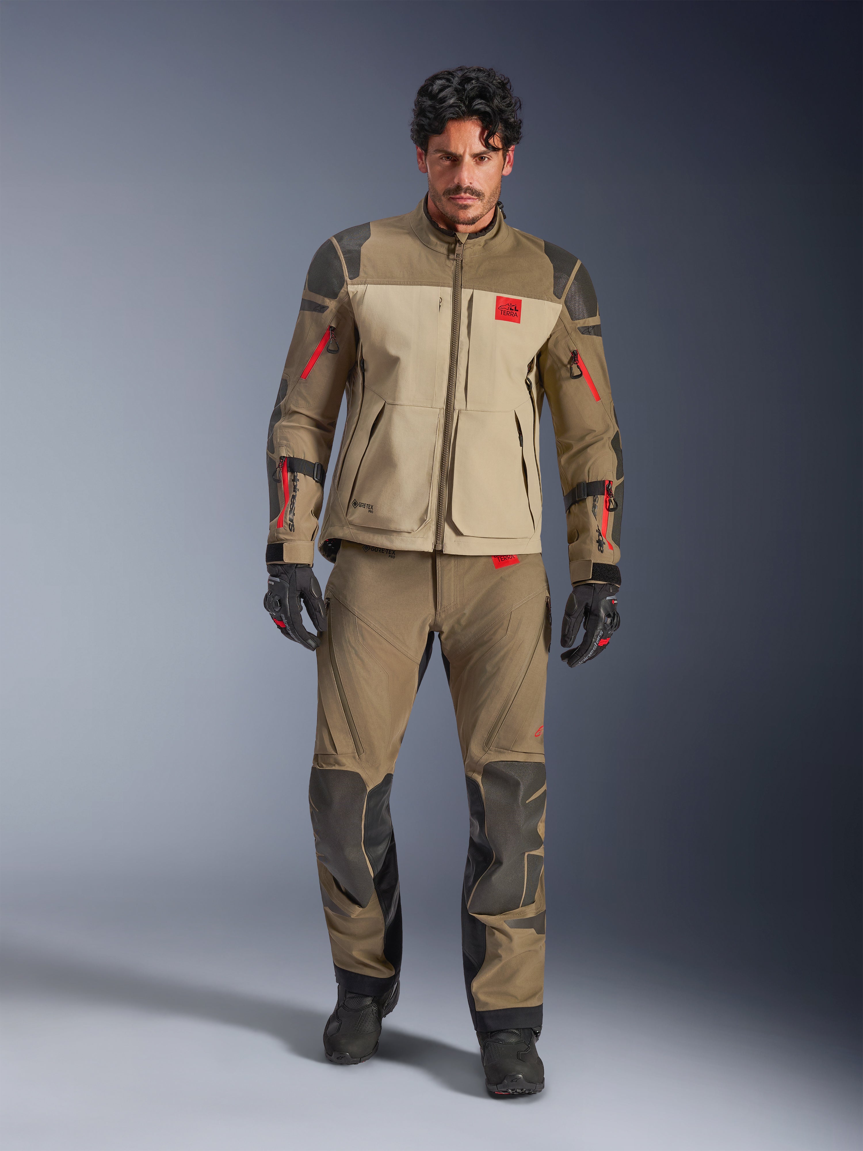 Alpinestars Nazca 3L GORE-TEX PRO Jacke, Walnut Rock Brown, Adventure-Motorradjacke getragen mit passender Hose und Handschuhen, rote Reißverschluss-Akzente, „All Terra“-Brustlogo, robuste Textilkonstruktion.