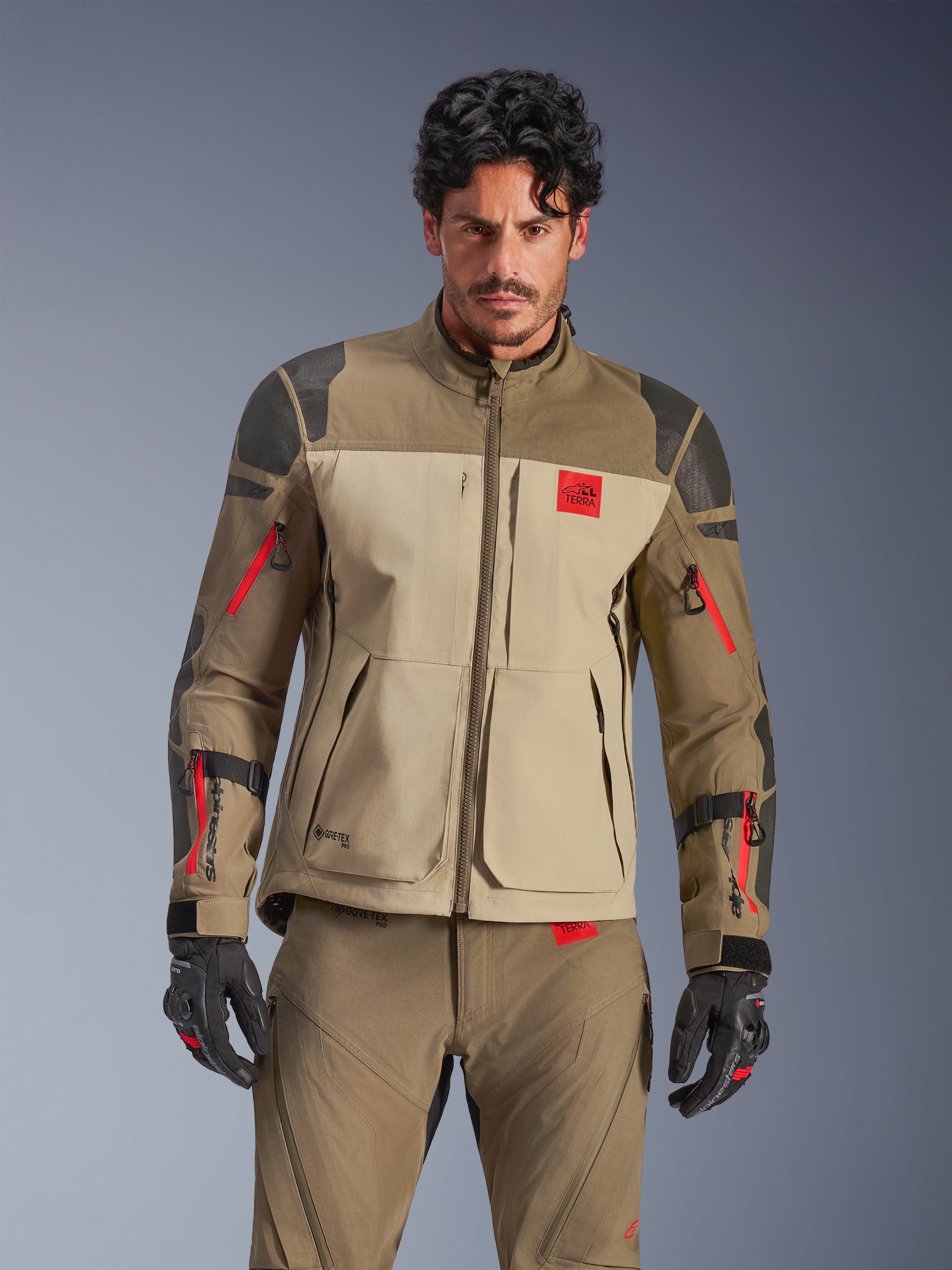 Alpinestars Nazca 3L GORE-TEX PRO Jacke, Adventure-Motorrad-Touringjacke, Walnut Rock Brown und Beige mit roten Akzenten, schwarze Superfabric-Schulterverstärkungen, rote Reißverschlüsse an den Ärmeln, getragen von einem Fahrer mit passender Ausrüstung, hochwertige technische Adventure-Bekleidung