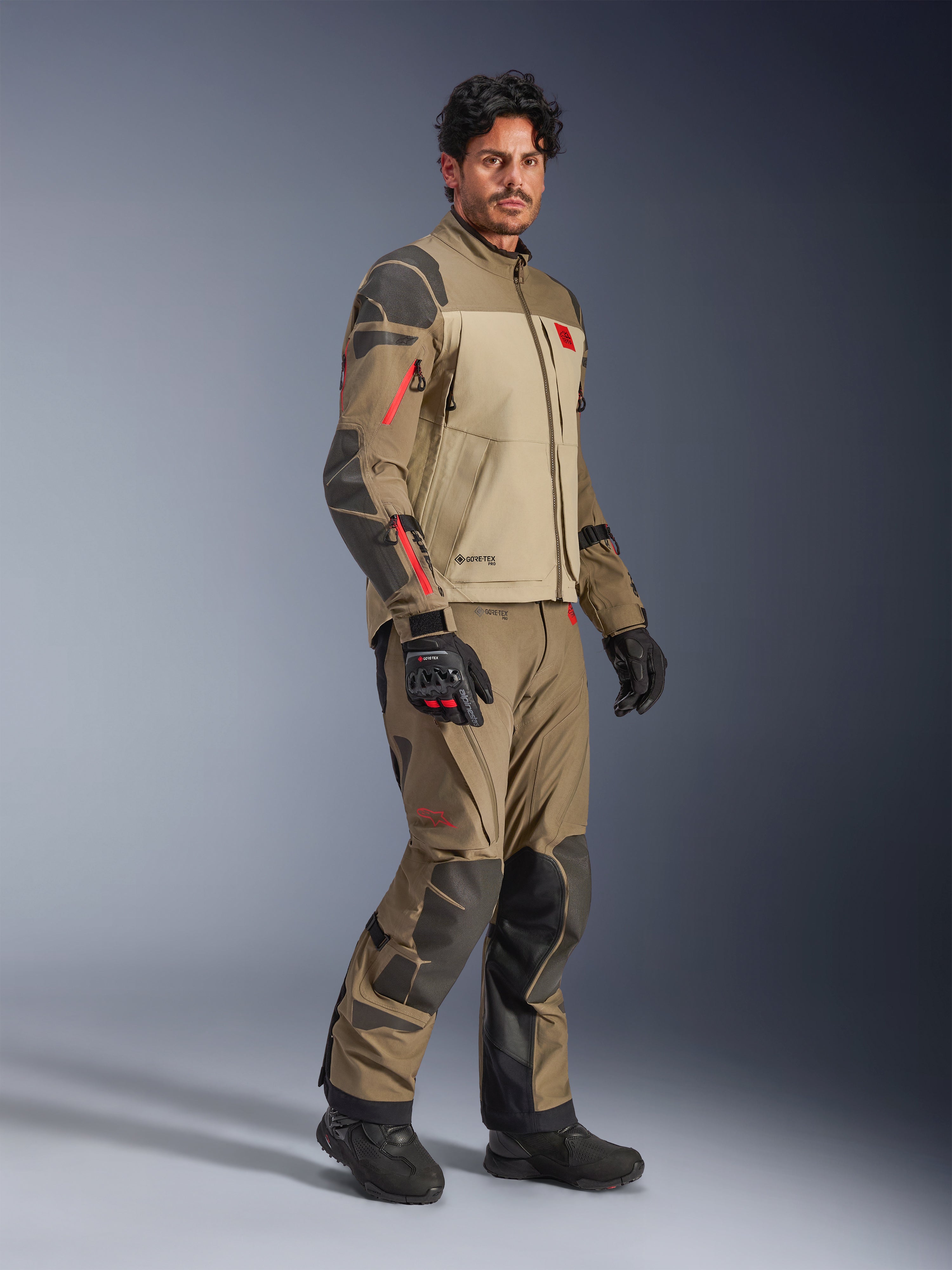 Alpinestars Nazca 3L GORE-TEX PRO Jacke, Walnut Rock Brown, Adventure-Motorradjacke getragen mit passender Hose und schwarzen Handschuhen, mit dunkelgrauen Verstärkungen, roten Reißverschluss-Details und einer hochleistungsfähigen laminierten Außenschicht.
