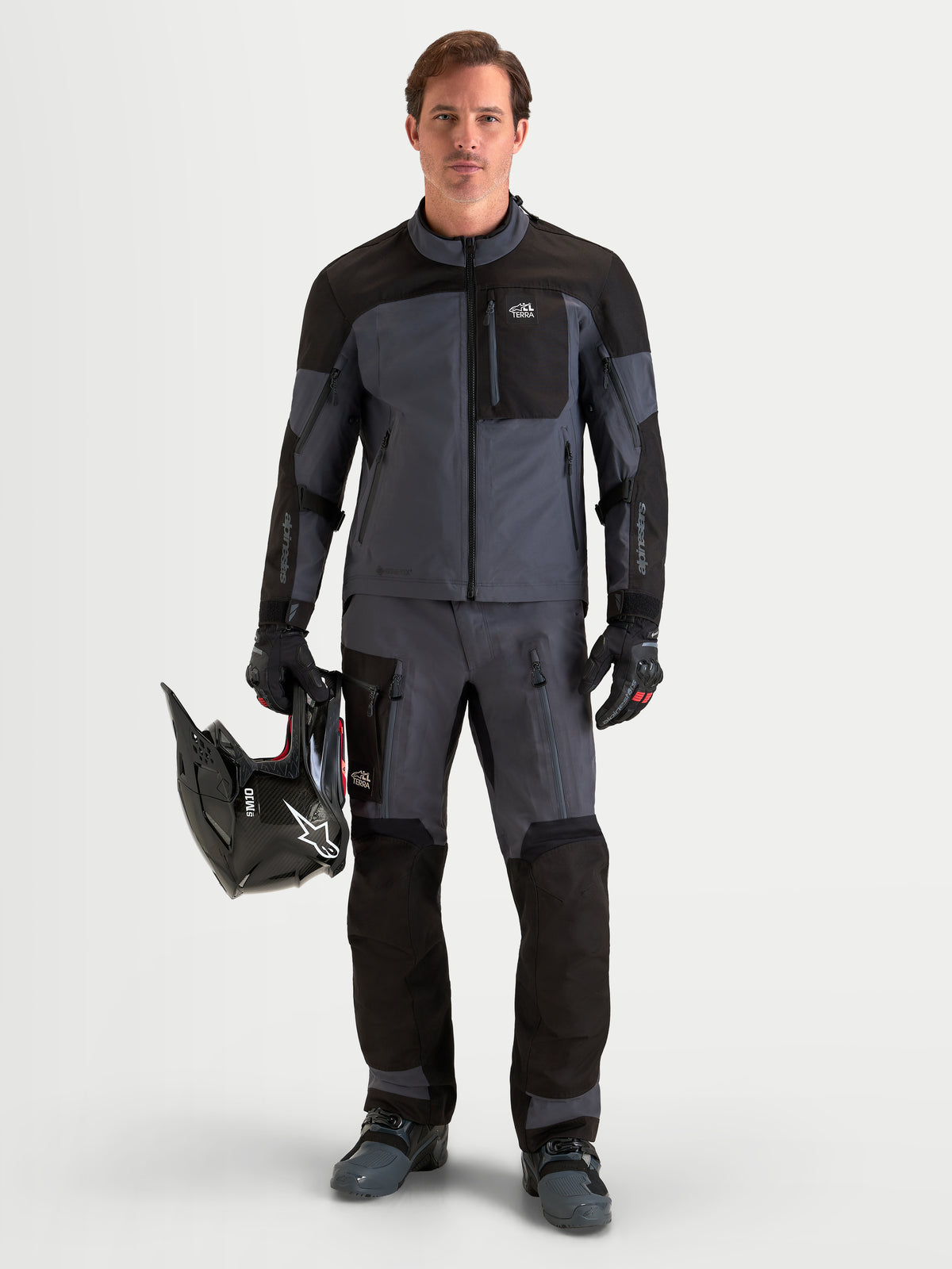 Alpinestars Arenis Gore-Tex Jacke, Herren-Adventure-Motorradjacke in Schwarz und Dunkelgrau, getragen von einem Fahrer, der einen Helm hält, mit wasserdichter GORE-TEX-Textilkonstruktion