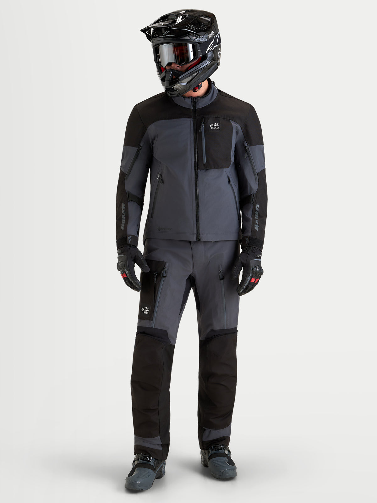 Alpinestars Arenis Gore-Tex Jacke, Herren-Adventure-Motorradjacke in Schwarz und Dunkelgrau, getragen von einem Fahrer mit passendem Helm und Schutzausrüstung, Ganzkörperansicht von vorne.
