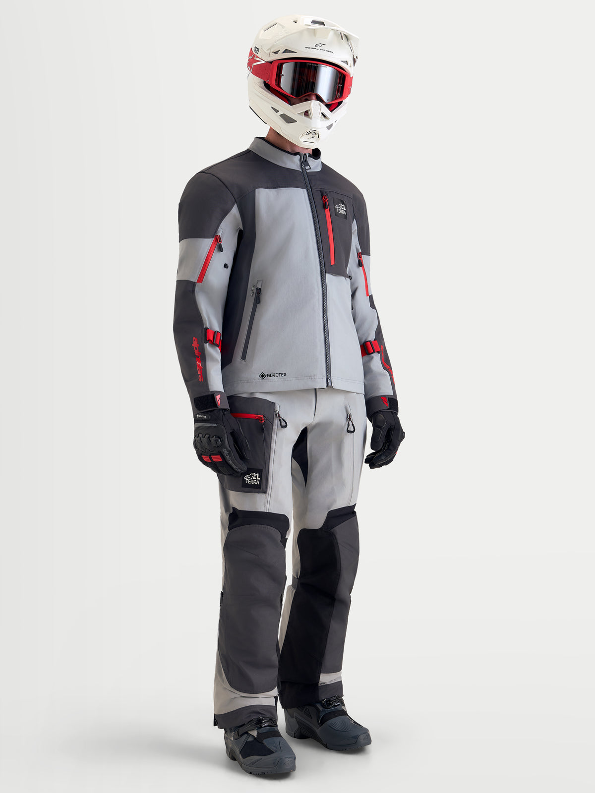Alpinestars Arenis Gore-Tex Jacke, Herren-Adventure-Motorradjacke in Mittelgrau, Dunkelgrau und Schwarz mit roten Akzenten, vollständige Schutzausrüstung mit passender Hose, Helm und Schutzbrille an Model.