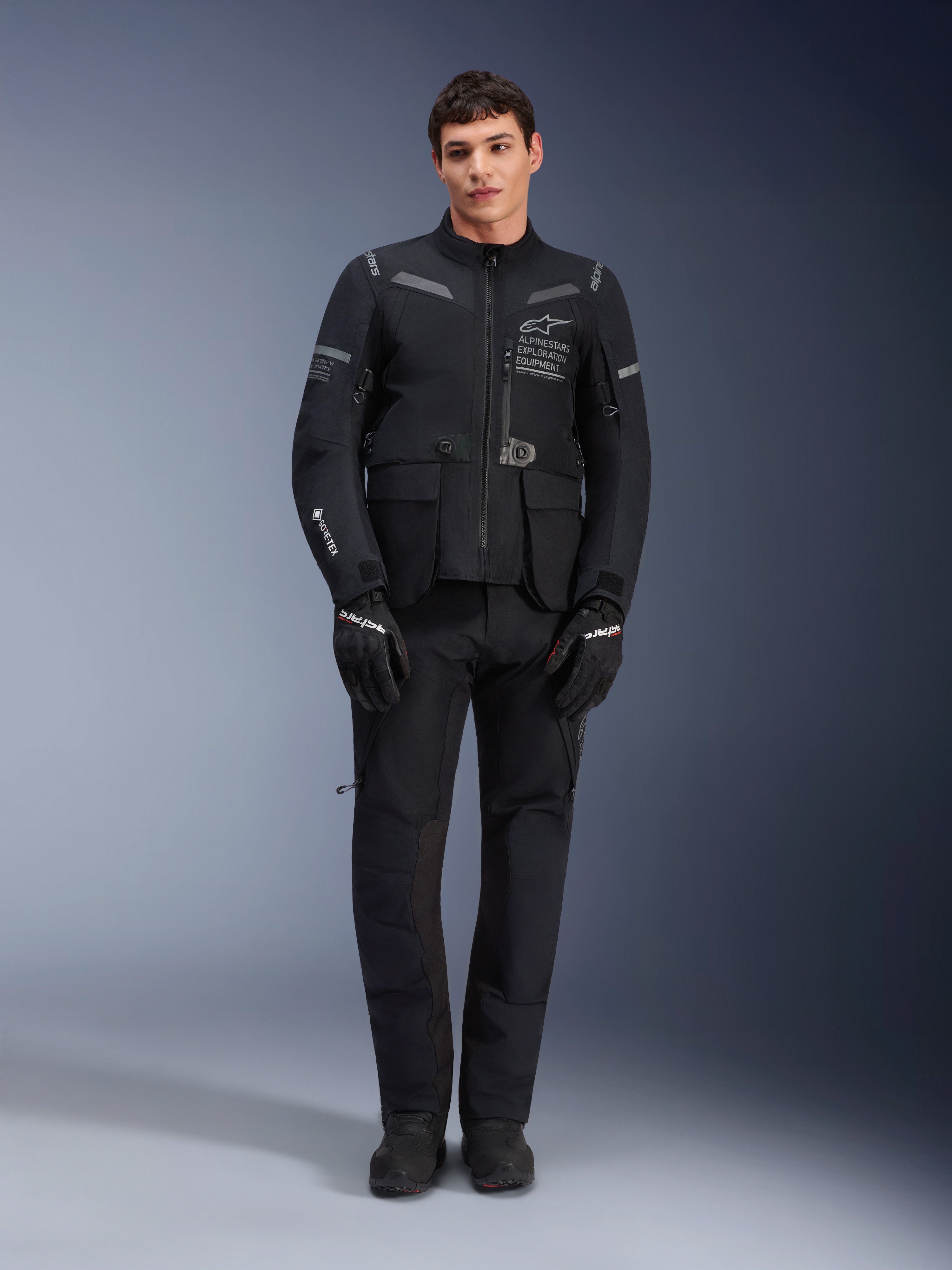 Alpinestars ST-7 2L GORE-TEX Jacke, Adventure-Touring-Motorradjacke, Schwarz und Dunkelgrau, getragen mit passender schwarzer Motorradhose und Handschuhen, mit mehreren Cargotaschen, reflektierenden Akzenten und Alpinestars Exploration Equipment-Branding auf der Brust, funktionales Design für Straßen- und Geländefahrten