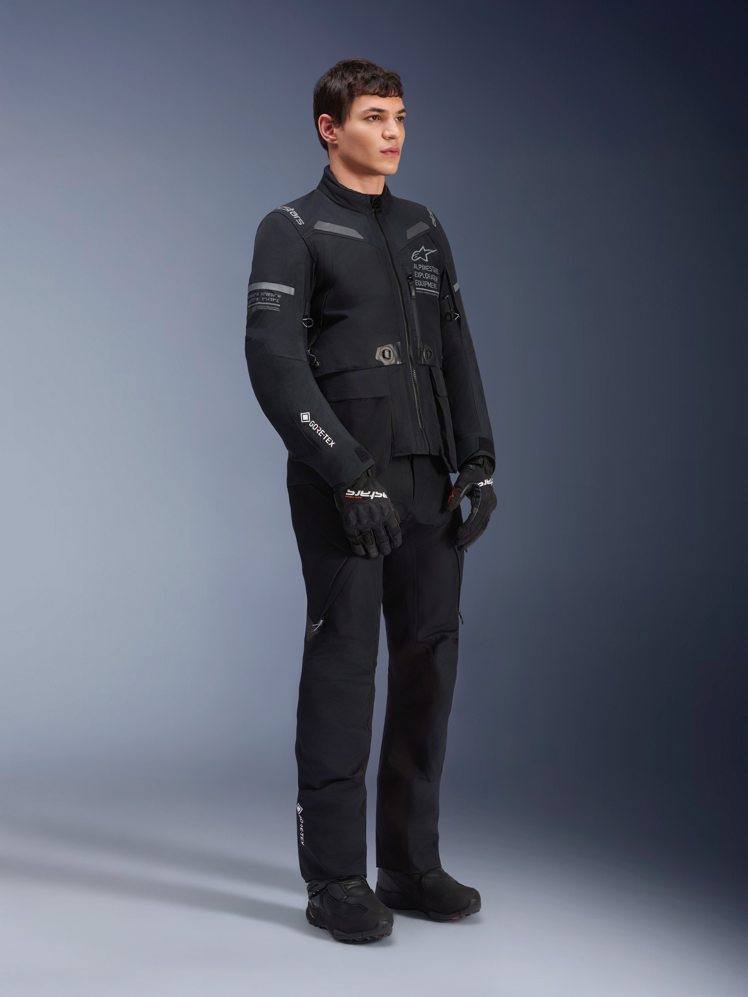 Männliches Model trägt Alpinestars ST-7 2L GORE-TEX Jacke in Black Dark Gray, Adventure-Touring-Motorradjacke, passende schwarze Hose und Handschuhe, Dreiviertel-Ganzkörperansicht, mit GORE-TEX Logo und Alpinestars Exploration Equipment Branding