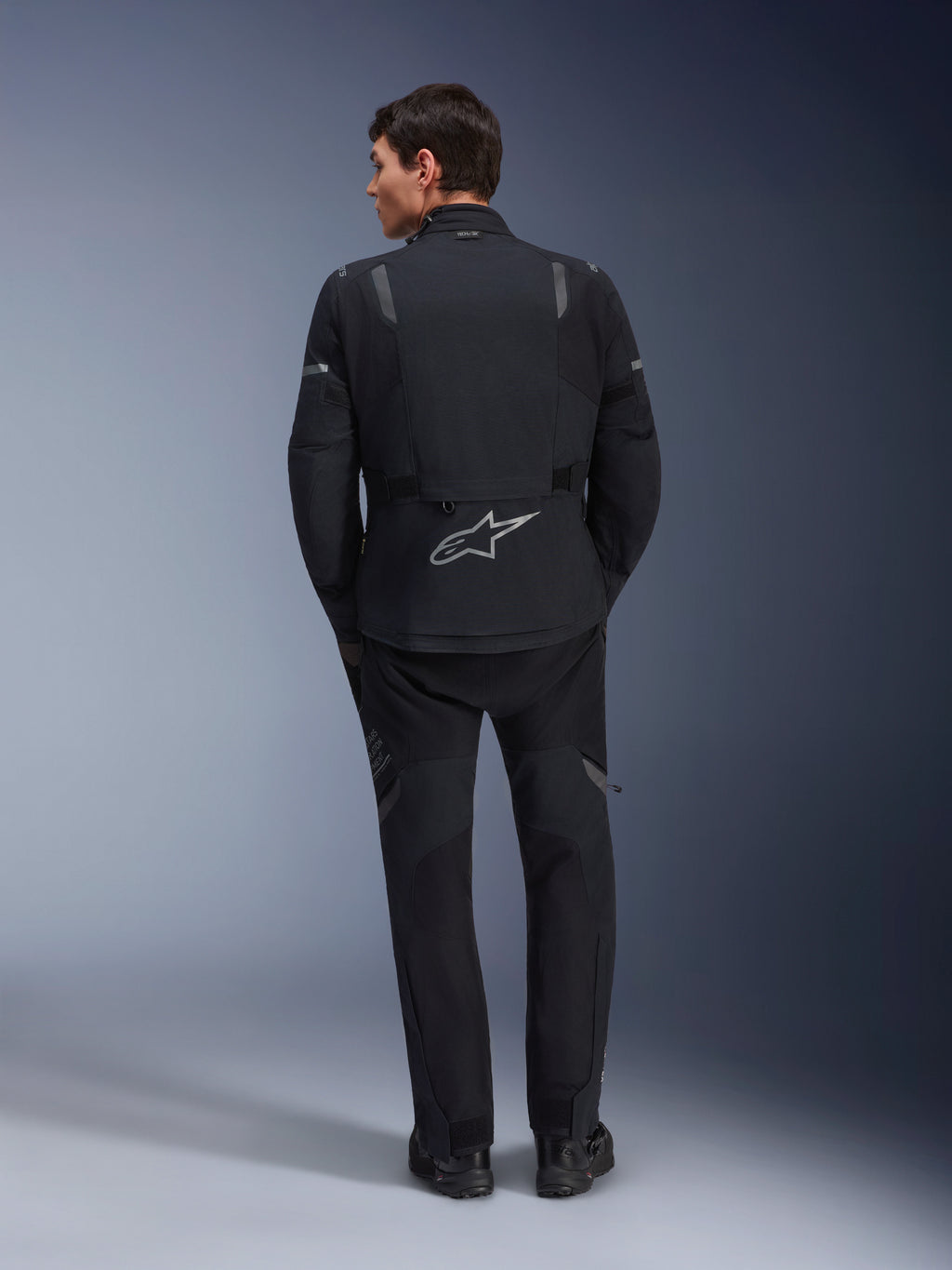 Alpinestars ST-7 2L GORE-TEX Jacke, Adventure-Motorradjacke, Schwarz und Dunkelgrau, Rückansicht eines Mannes in Jacke und passender schwarzer Hose, großes reflektierendes Astar-Logo auf dem unteren Rücken, wasserdichte Textilausrüstung für Touren und Straßenfahrten
