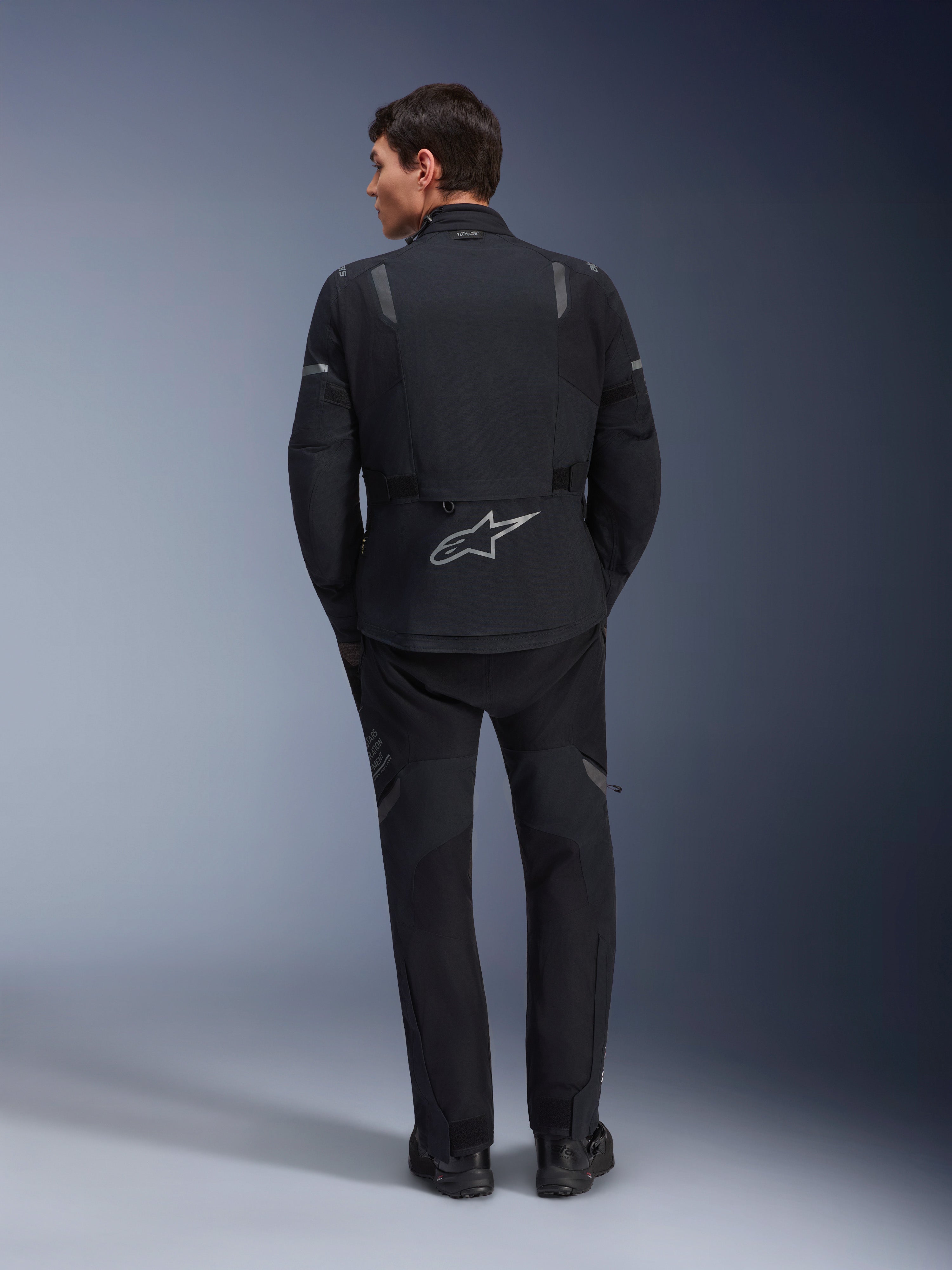 Alpinestars ST-7 2L GORE-TEX Jacke, Adventure-Motorradjacke, Schwarz und Dunkelgrau, Rückansicht eines Mannes in Jacke und passender schwarzer Hose, großes reflektierendes Astar-Logo auf dem unteren Rücken, wasserdichte Textilausrüstung für Touren und Straßenfahrten