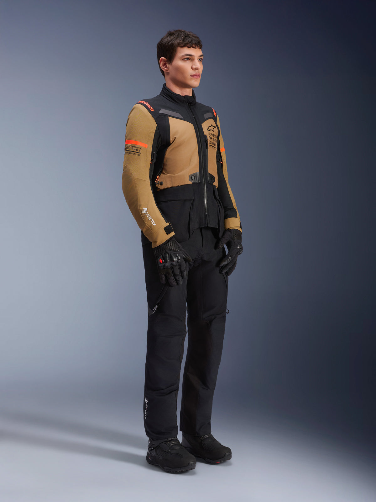 Alpinestars ST-7 2L GORE-TEX Jacke, Motorradjacke, Gobi-Braun und Schwarz, getragen von einem Model mit passender schwarzer Hose und Handschuhen, mit braunen Einsätzen an Brust und Ärmeln, schwarzen technischen Akzenten und orangefarbenem Branding, Adventure-Touring-Motorradausrüstung.
