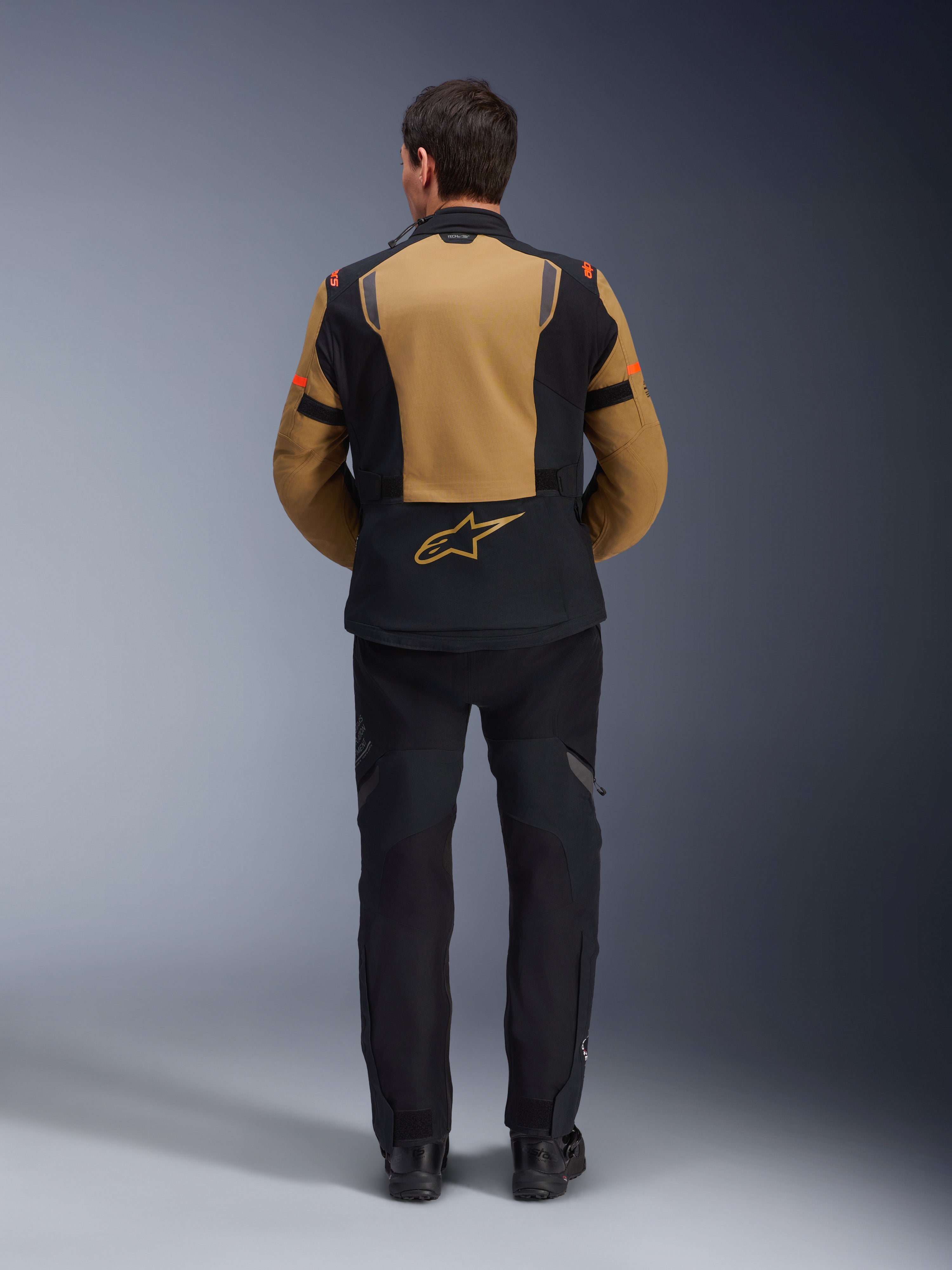 Alpinestars ST-7 2L GORE-TEX Jacke, Adventure-Motorradjacke, Gobi-Braun und Schwarz, Rückansicht mit ergonomischen Einsätzen und Alpinestars-Logo, getragen mit schwarzer Motorradhose und Stiefeln, wasserdichte Tourenbekleidung