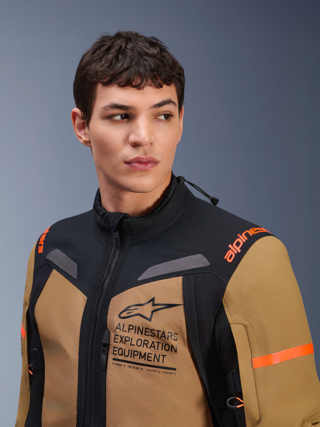 Alpinestars ST-7 2L GORE-TEX Jacke, Gobi-Braun und Schwarz, getragen von einem männlichen Model in einer Studioumgebung, mit markantem 'ALPINESTARS EXPLORATION EQUIPMENT' Brust-Branding, orangefarbenen Schulterlogos und technischer wasserdichter Textilkonstruktion für Adventure-Fahrten.