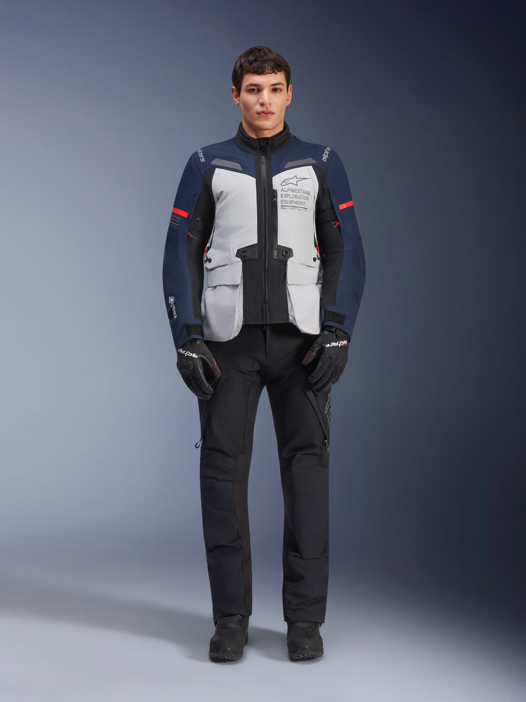 Alpinestars ST-7 2L GORE-TEX Jacke, Adventure-Touren-Motorradjacke, Eisgrau, Dunkelblau und Schwarz, getragen mit schwarzer Hose und Handschuhen, große Cargo-Taschen vorne, rote Akzente an den Ärmeln, laminierte GORE-TEX-Außenschicht für wasserdichte Performance.