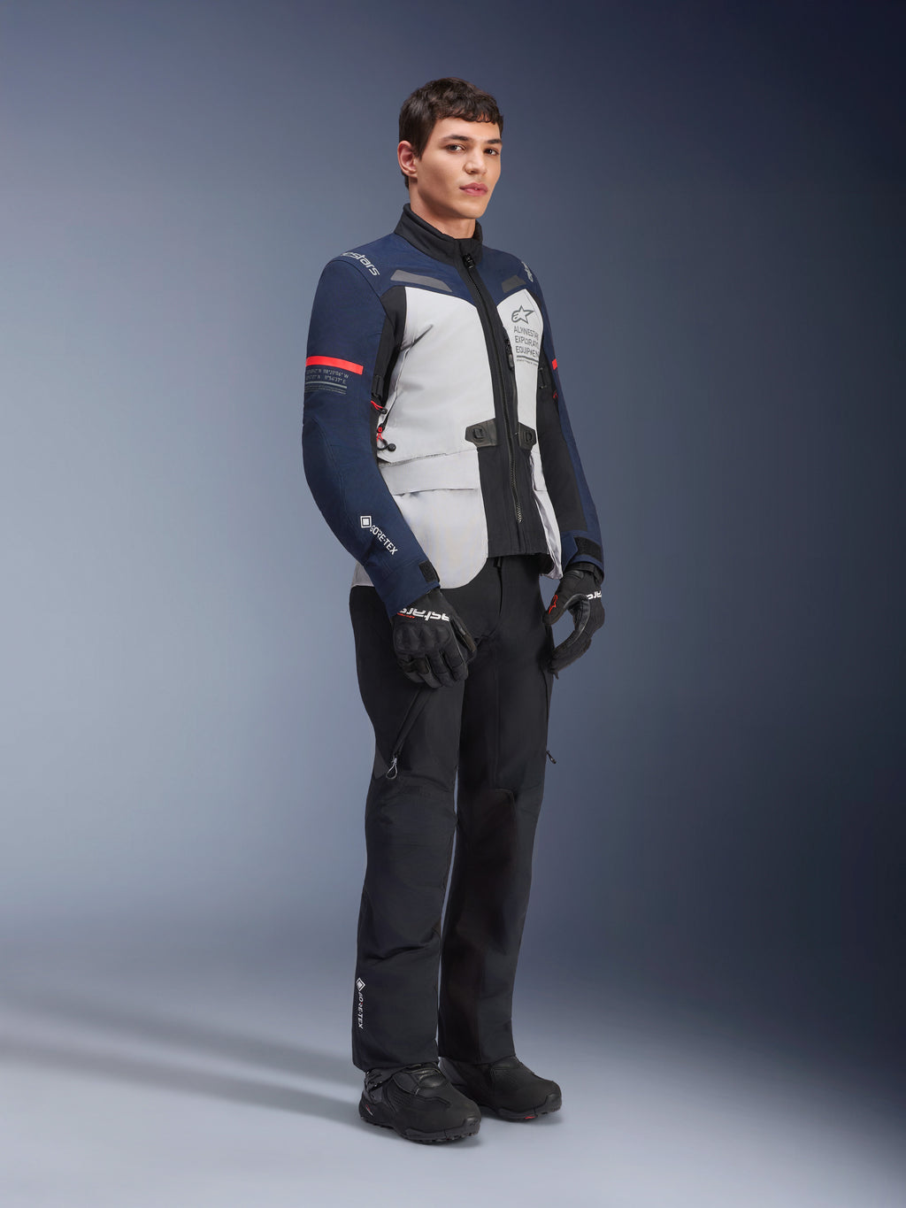 Alpinestars ST-7 2L GORE-TEX Jacke in Ice Gray, Dunkelblau und Schwarz mit roten Akzenten, getragen von einem männlichen Modell mit schwarzer Motorradhose und Handschuhen, professionelle Adventure-Touring-Motorradausrüstung