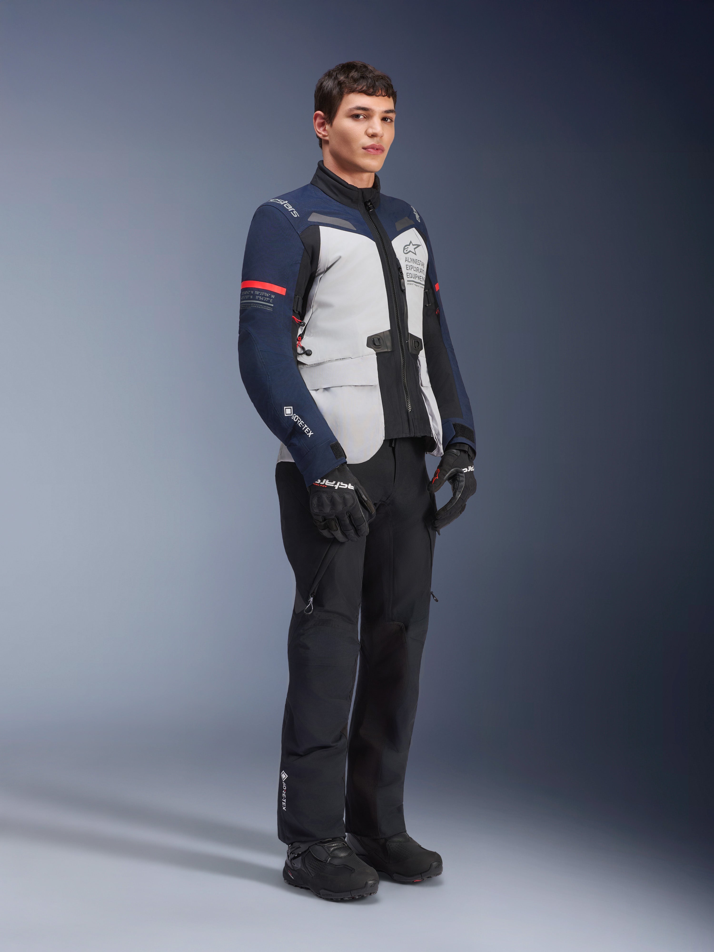 Alpinestars ST-7 2L GORE-TEX Jacke in Ice Gray, Dunkelblau und Schwarz mit roten Akzenten, getragen von einem männlichen Modell mit schwarzer Motorradhose und Handschuhen, professionelle Adventure-Touring-Motorradausrüstung