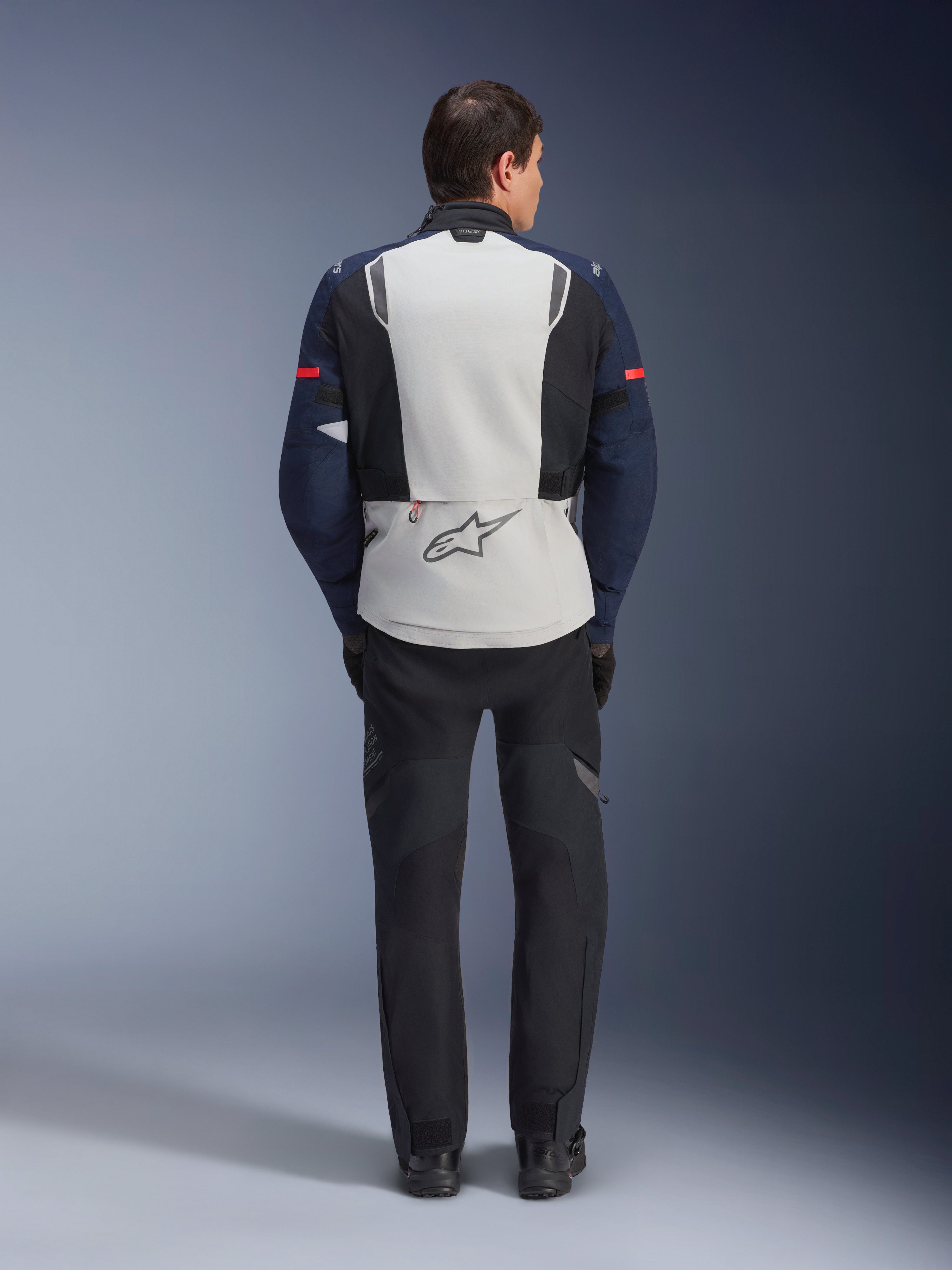 Alpinestars ST-7 2L GORE-TEX Jacke, Rückansicht eines Fahrers in einer Adventure-Touren-Motorradjacke in Ice Gray, Dunkelblau und Schwarz, mit großem Rückenpanel und Alpinestars-Logo sowie passender schwarzer Hose, konzipiert für Fahrten bei jedem Wetter.