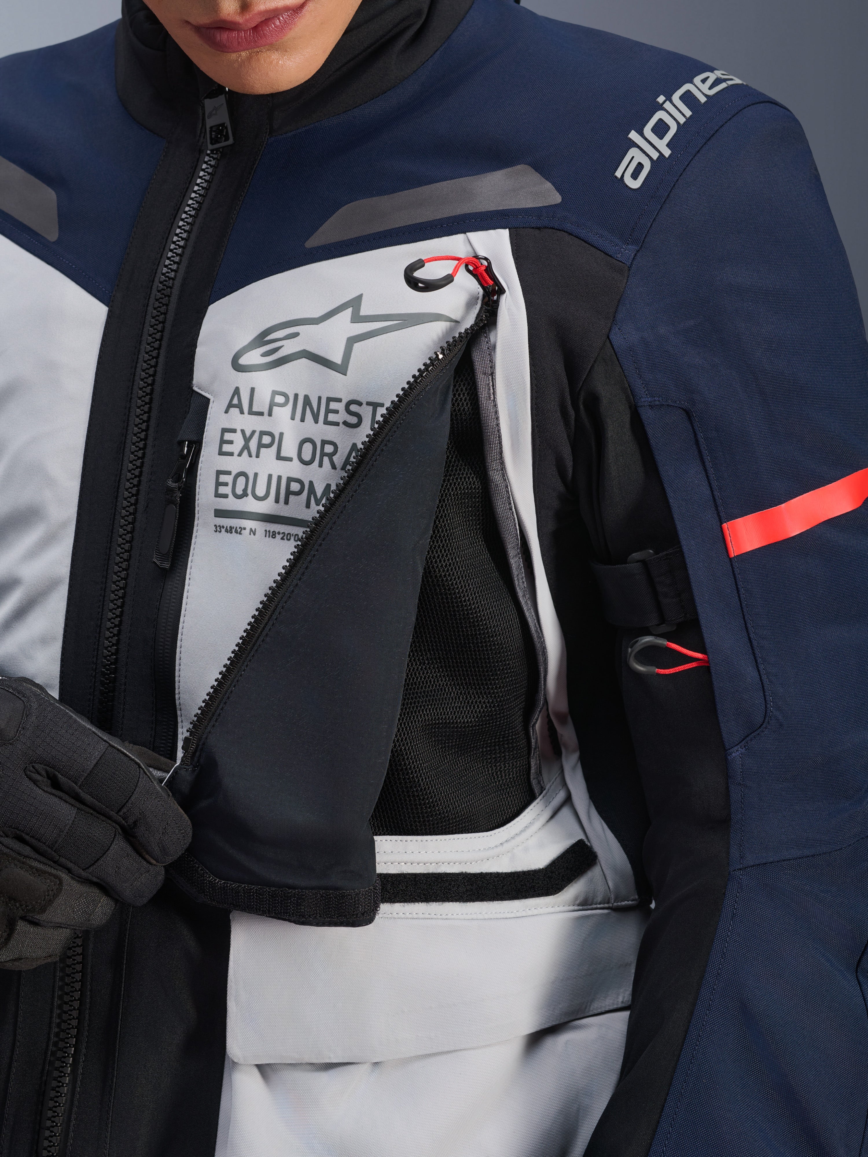 Alpinestars ST-7 2L GORE-TEX Jacke, Adventure-Tourenjacke, Ice Gray, Dunkelblau und Schwarz, Detailansicht des geöffneten Brust-Belüftungspanels mit schwarzem Mesh, Alpinestars Exploration Equipment Logo und roten Akzenten an den Zuglaschen