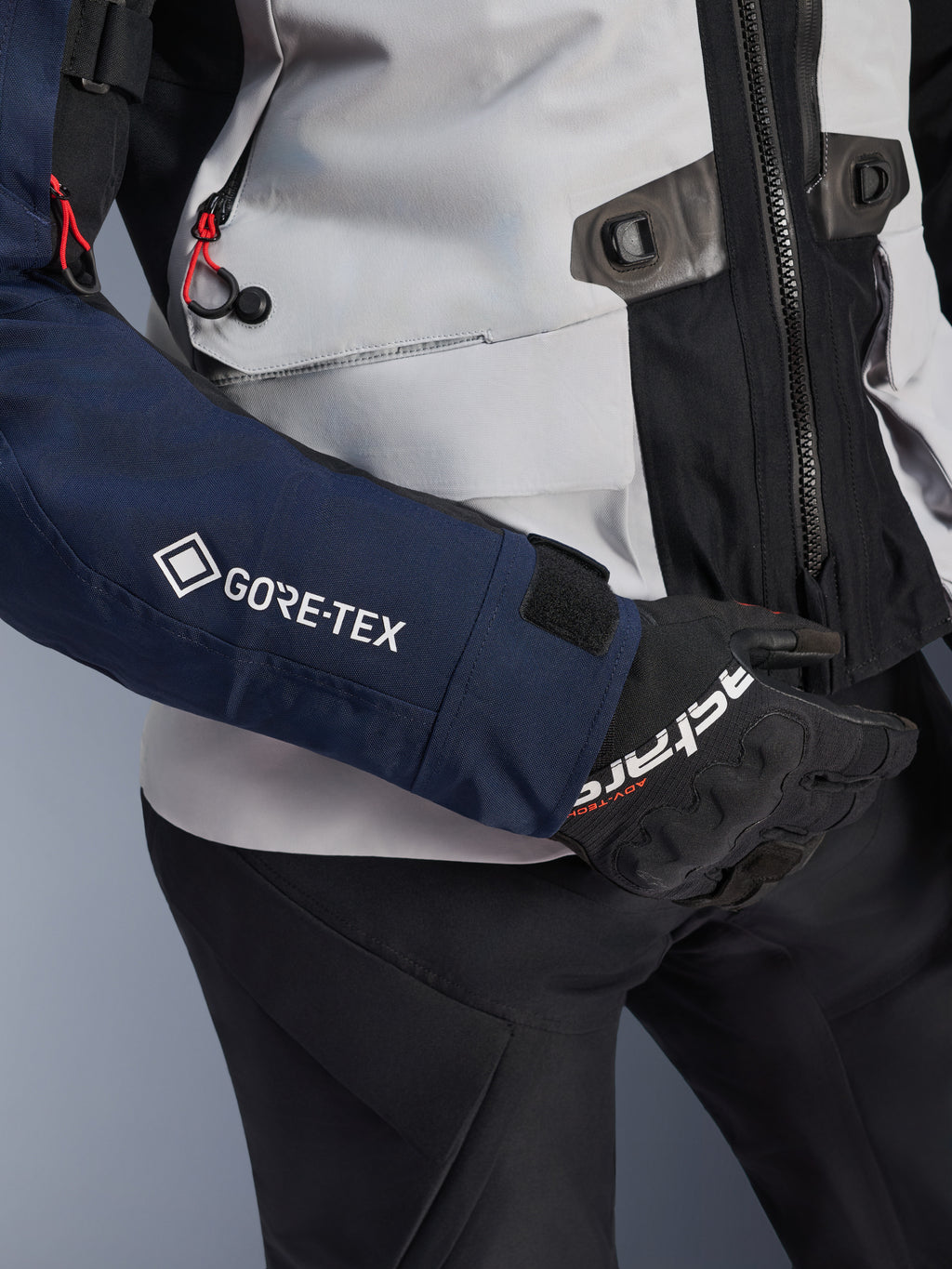Alpinestars ST-7 2L GORE-TEX Jacke, Nahaufnahme eines dunkelblauen Ärmels mit weißem GORE-TEX Logo, eisgrauer Torso mit schwarzen Details und roten Kordelakzenten, kombiniert mit schwarzen Alpinestars Handschuhen und Hose, Adventure-Touring-Motorradausrüstung