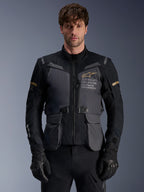 Alpinestars ST-7 2L GORE-TEX Jacke, Herren Adventure-Touring-Motorradjacke, Dunkelgrau und Schwarz mit khakifarbenen Akzenten, mit „Alpinestars Exploration Equipment“-Branding, wasserdichten Cargotaschen und hohem Kragen, getragen von einem Model mit schwarzen Handschuhen