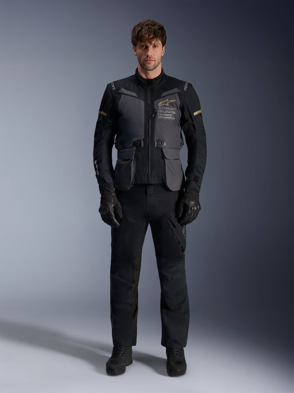 Männliches Model trägt die Alpinestars ST-7 2L GORE-TEX Jacke in Deep Gray Black Dark Khaki, mit dem „ALPINESTARS EXPLORATION EQUIPMENT“-Branding auf der Brust, kombiniert mit passender schwarzer Motorradhose, Handschuhen und Stiefeln, Studio-Frontansicht.