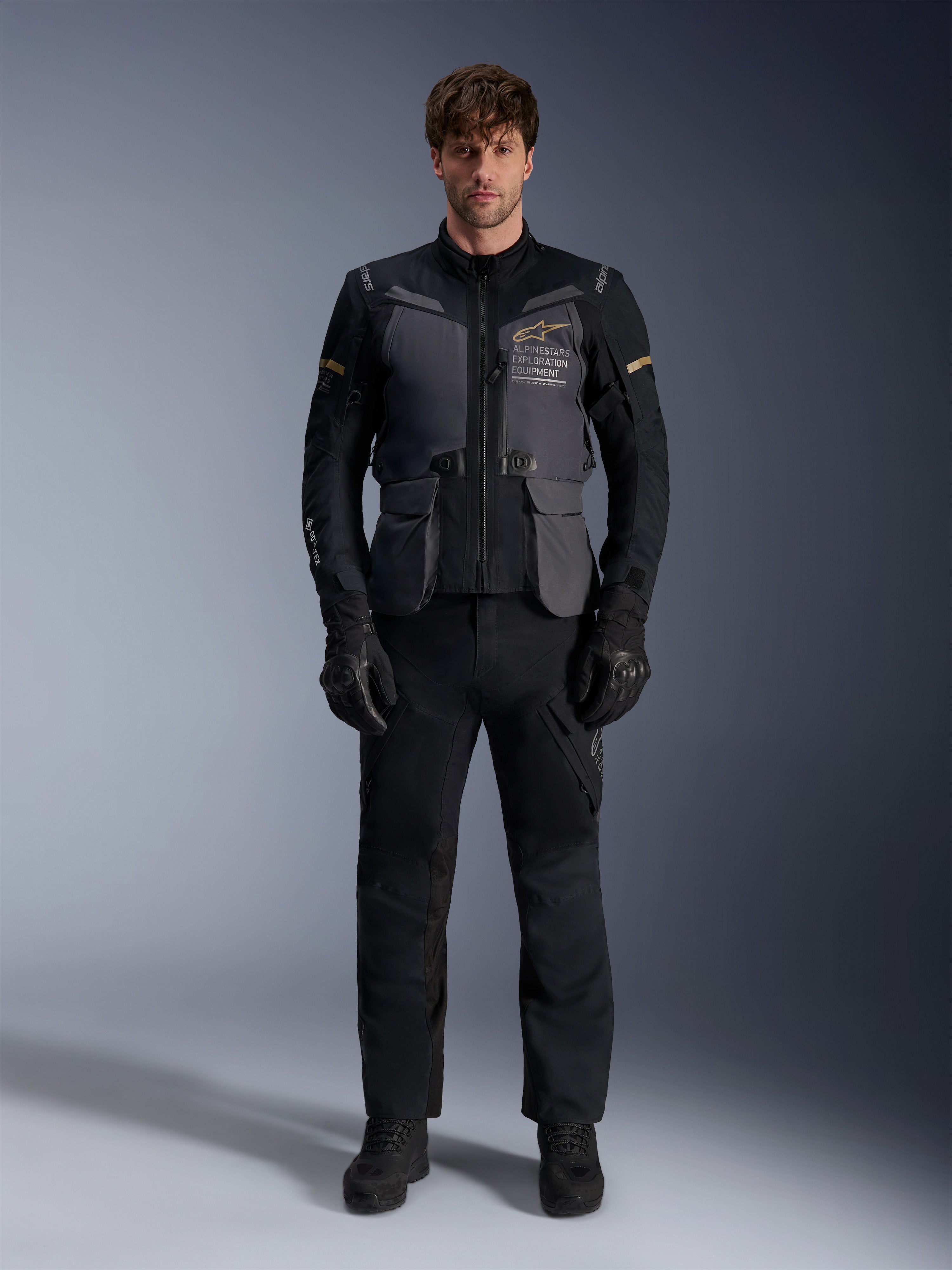 Männliches Model trägt die Alpinestars ST-7 2L GORE-TEX Jacke in Deep Gray Black Dark Khaki, mit dem „ALPINESTARS EXPLORATION EQUIPMENT“-Branding auf der Brust, kombiniert mit passender schwarzer Motorradhose, Handschuhen und Stiefeln, Studio-Frontansicht.