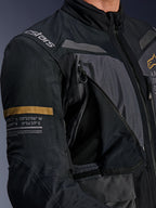 Alpinestars ST-7 2L GORE-TEX Jacke, Dunkelgrau und Schwarz, Nahaufnahme mit geöffnetem Belüftungspanel mit Reißverschluss und Mesh an der Brust, Alpinestars-Logo auf der Schulter und khakifarbenem Streifen auf dem Ärmel mit aufgedruckten geografischen Koordinaten, technische Adventure-Touring-Motorradausrüstung