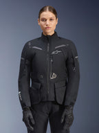 Frau in Alpinestars Stella ST-7 2L GORE-TEX Jacke, Schwarz mit dunkelgrauen Akzenten, Motorrad-Tourenjacke mit großen Cargo-Taschen und hohem Kragen, kombiniert mit schwarzen Handschuhen und Hosen, wasserdichte Adventure-Fahrbekleidung