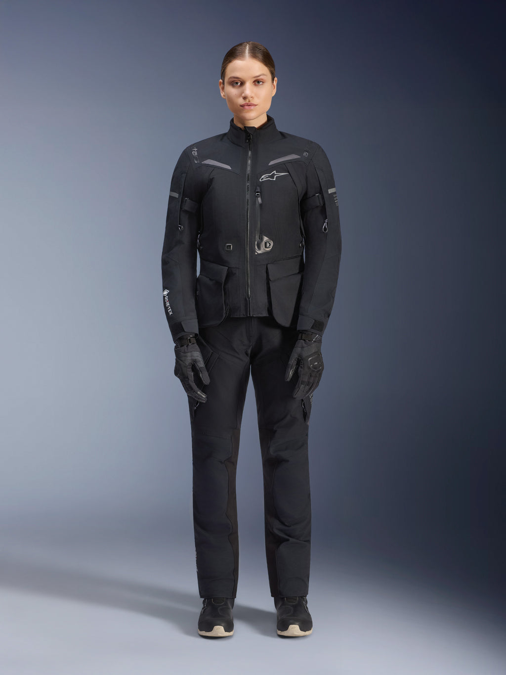 Motorradfahrerin in Alpinestars Damen Stella ST-7 2L GORE-TEX Jacke und Hose, schwarzes Motorrad-Tourenoutfit, wasserdichte und atmungsaktive technische Ausrüstung, mit passenden schwarzen Handschuhen und Stiefeln, Ganzkörper-Schutzausrüstung mit reflektierenden Elementen, modernes technisches Design für Adventure-Riding