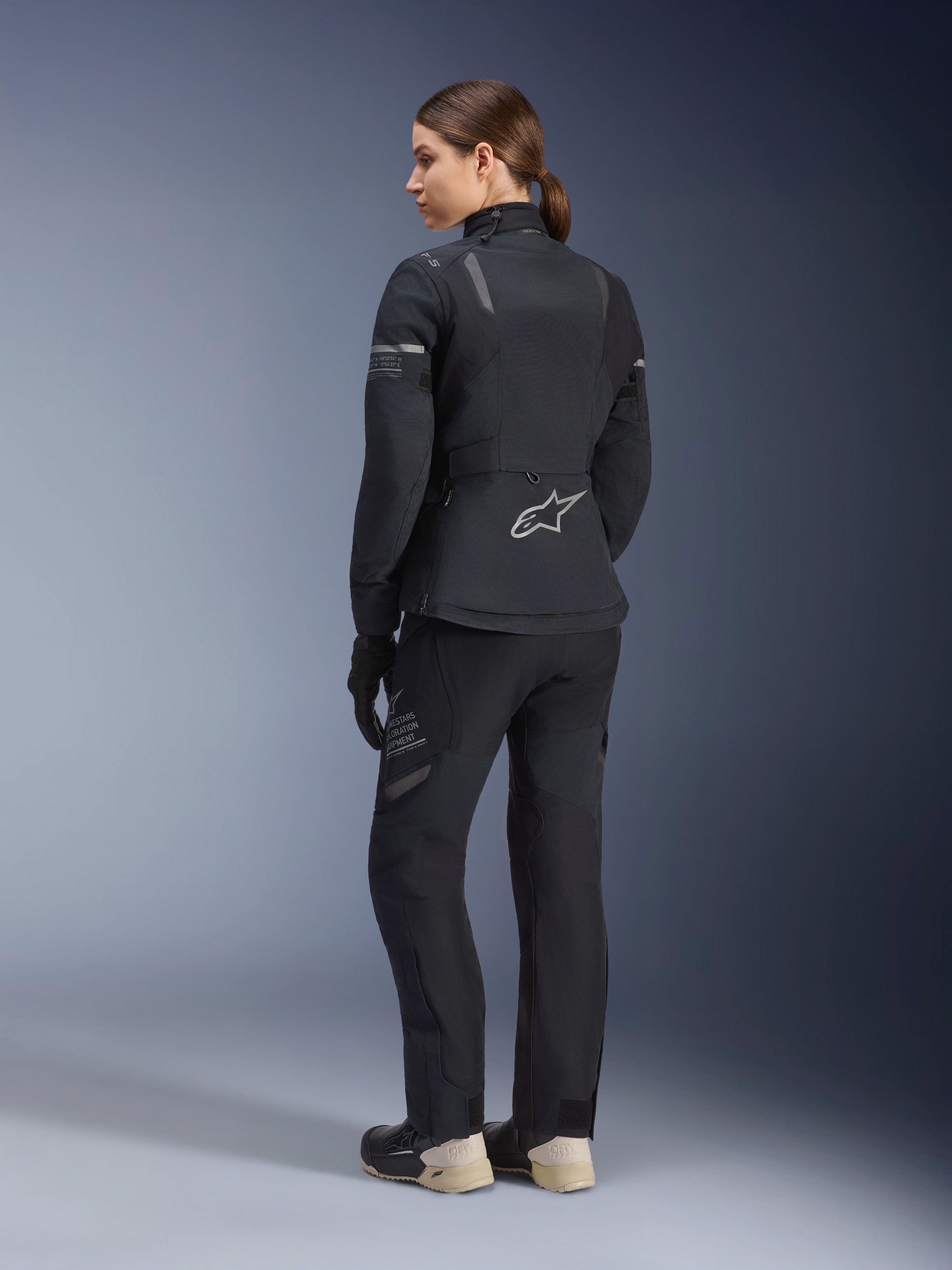 Stella ST-7 2L GORE-TEX Damenjacke, Schwarz und Dunkelgrau, Adventure-Tourenjacke in der Rückansicht, getragen mit passender schwarzer Hose und cremefarbenen Stiefeln, Alpinestars-Logo auf dem unteren Rücken, konzipiert für ganzjährige Straßentouren