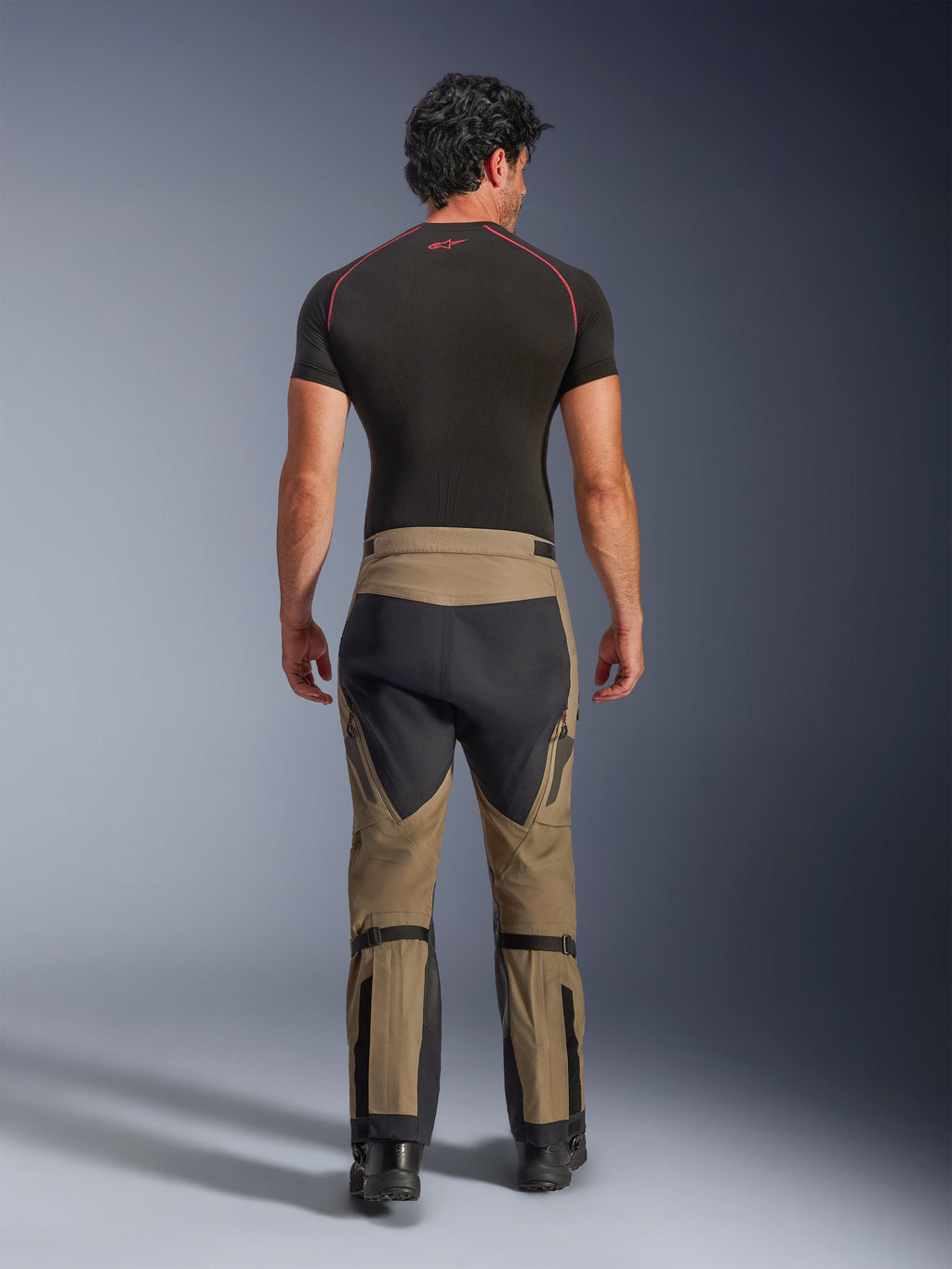 Alpinestars Nazca 3L GORE-TEX PRO Hose, Adventure-Motorradhose in Walnut Black, Rückansicht an einem Model mit schwarzem Base Layer und Stiefeln, zweifarbiges Design mit Wadenverstellern und Stretch-Paneelen.