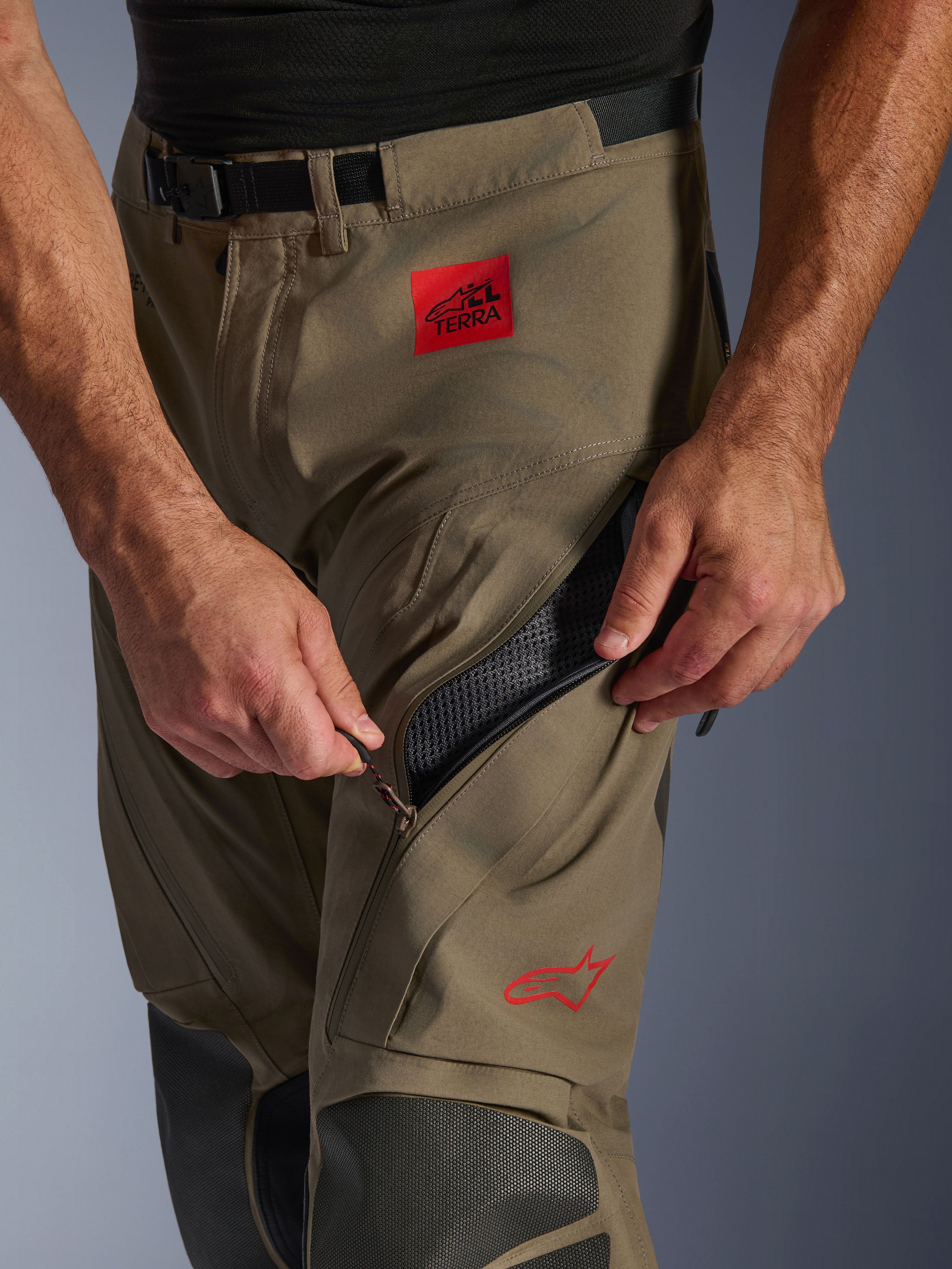 Alpinestars Nazca 3L GORE-TEX PRO Hose in Walnut Black, Nahaufnahme des Oberschenkelbereichs, eine Hand öffnet die große Belüftungsöffnung mit Reißverschluss, mit rotem ALL TERRA-Aufnäher und rotem Alpinestars-Logo, technische Adventure-Motorradausrüstung.